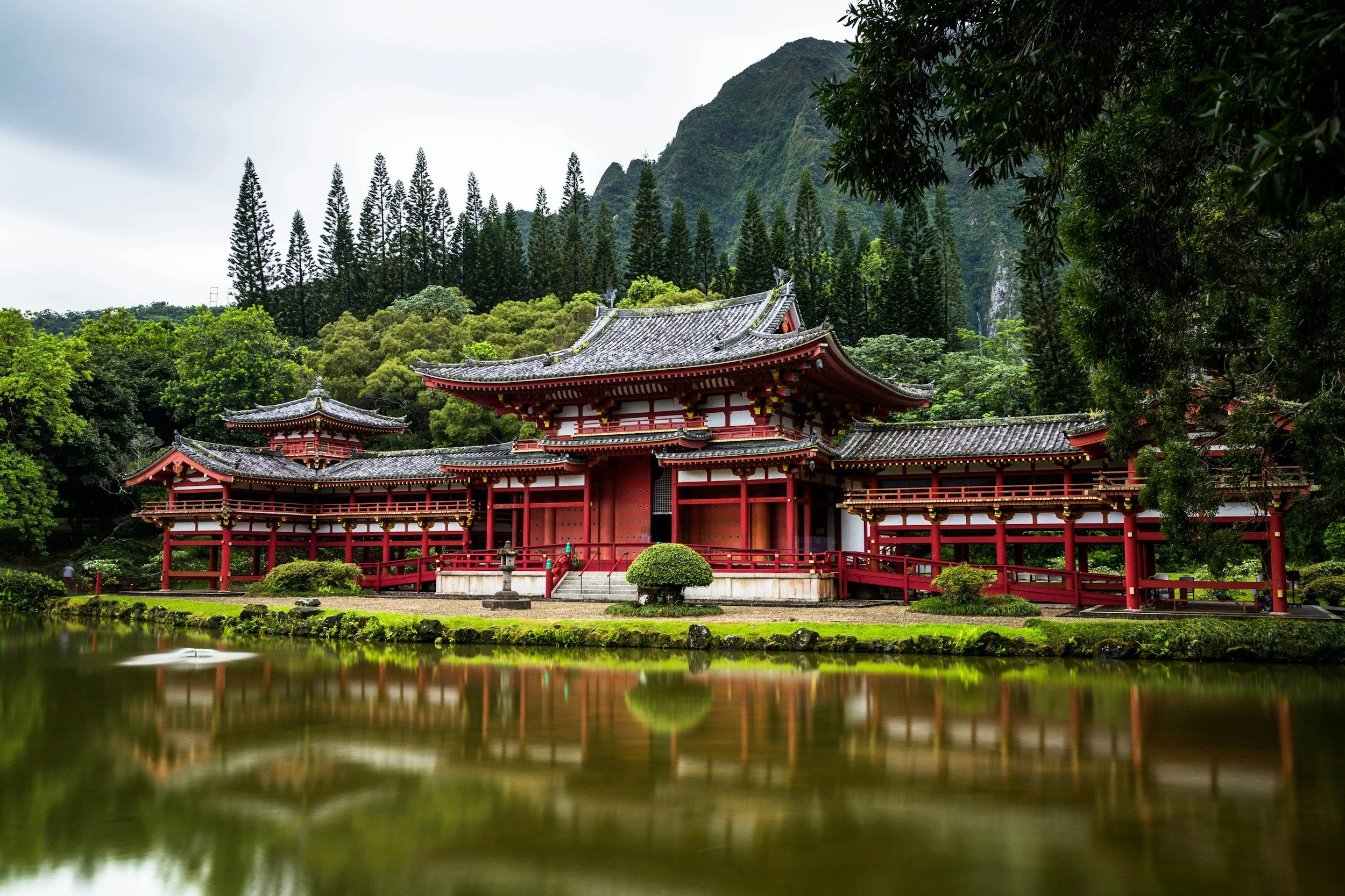 Traditional red and white Japanese temple surrounded by lush green trees and mountains, with a reflective pond in the foreground.