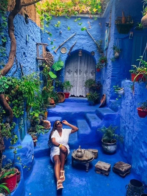 A woman sitting on blue stairs in a vibrant, flower-filled courtyard with blue walls, potted plants, and decorative items hanging on the walls.