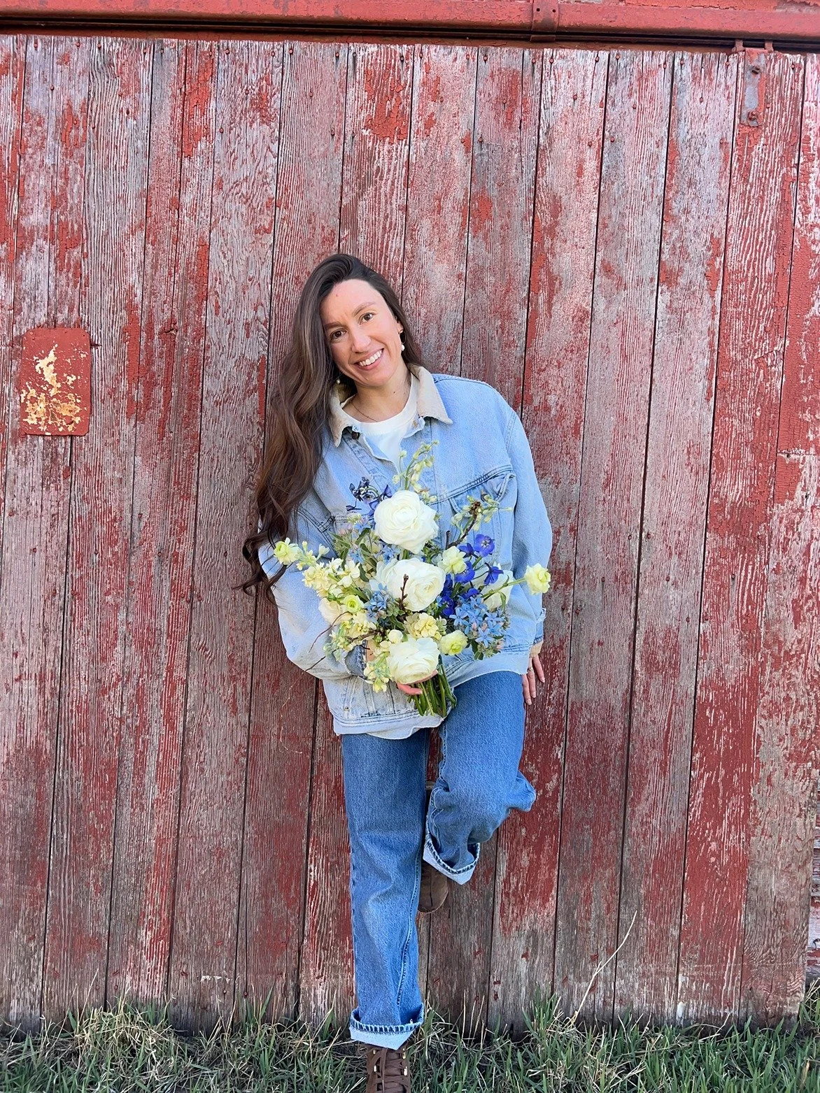 A woman smiling and holding a large bouquet of white, blue, and light yellow flowers, standing in front of a weathered red wooden wall.
