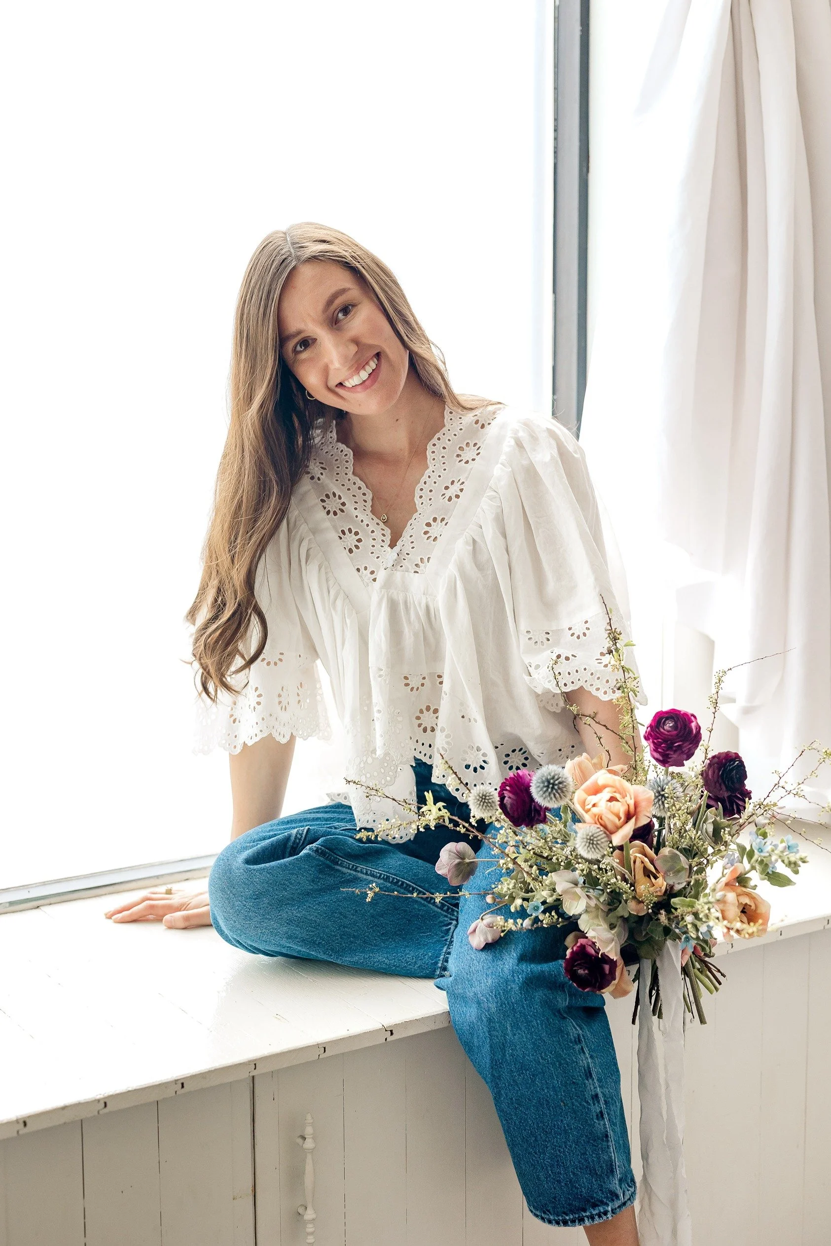 A woman with long wavy hair smiling while sitting on a white windowsill, holding a bouquet of pink, purple, and peach flowers.