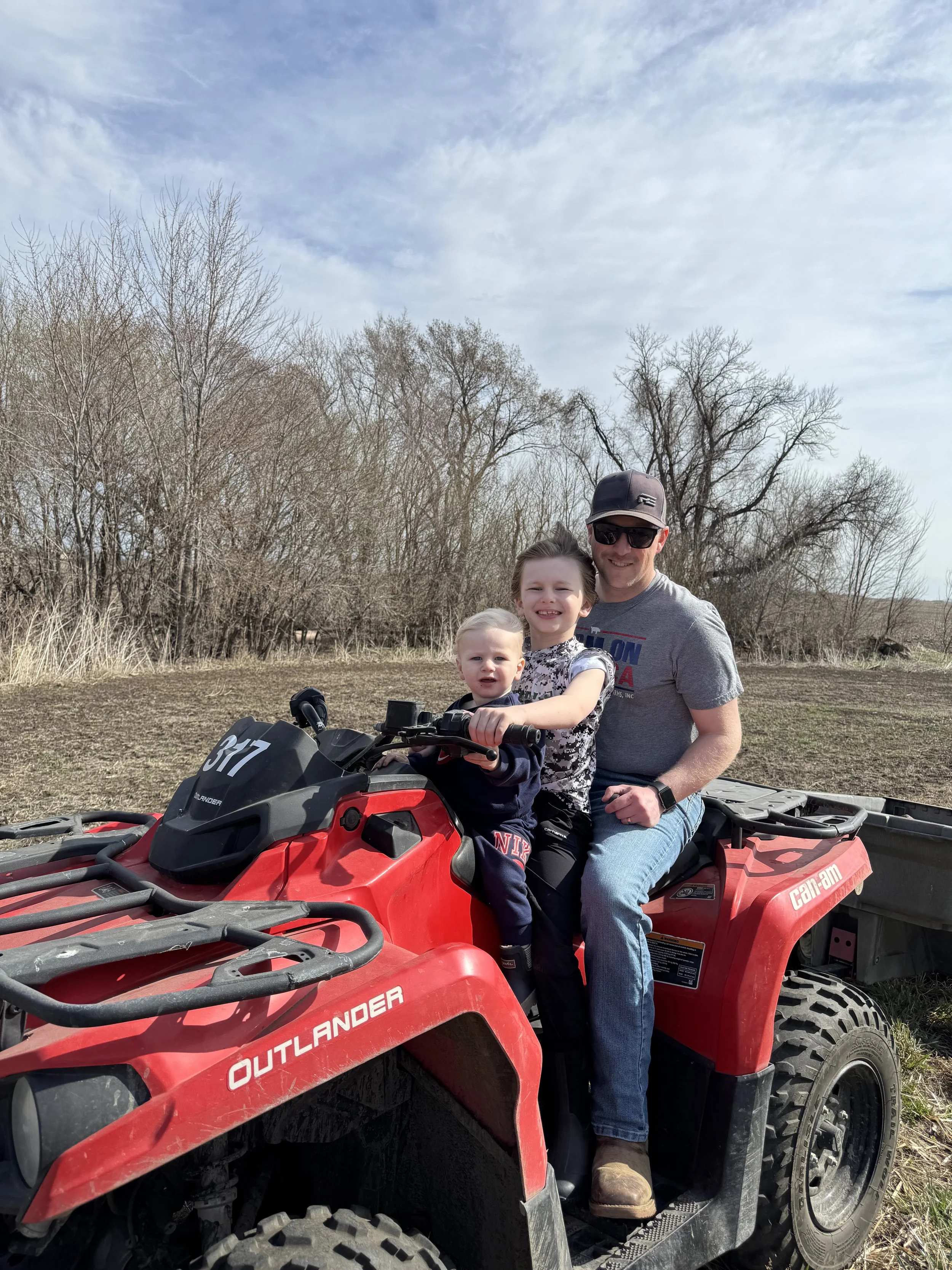 Three people working in a field on a farm or backyard, with a young boy sitting on an orange ATV and two men standing with shovels, a small green tractor in the background, and leafless trees under a partly cloudy sky.