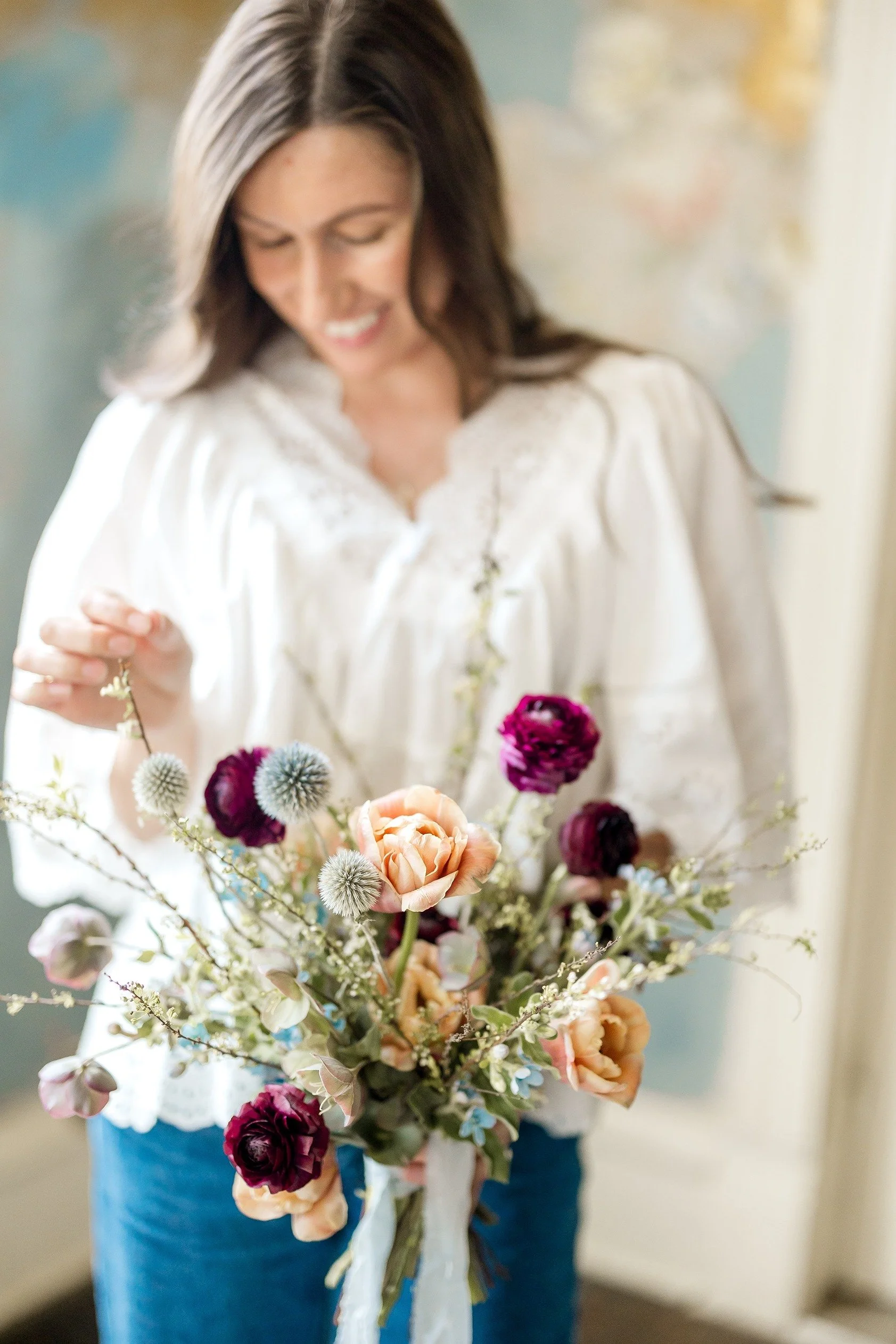 Woman in a white blouse holding a bouquet of colorful flowers, smiling and looking down.