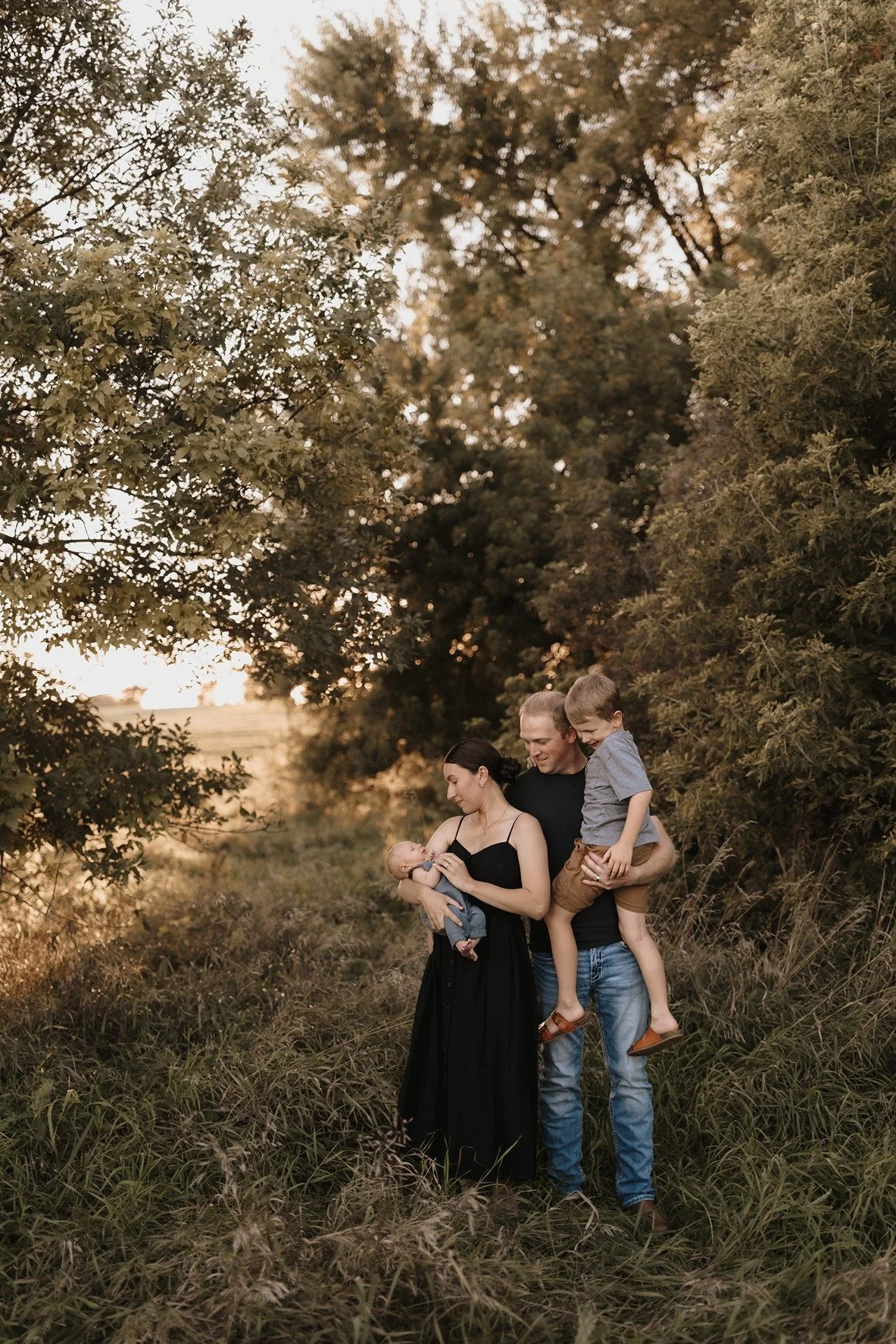 A family of four stands outdoors during sunset, with trees and tall grass around them. The woman holds a newborn baby, the man is holding an older child, and they are all smiling.