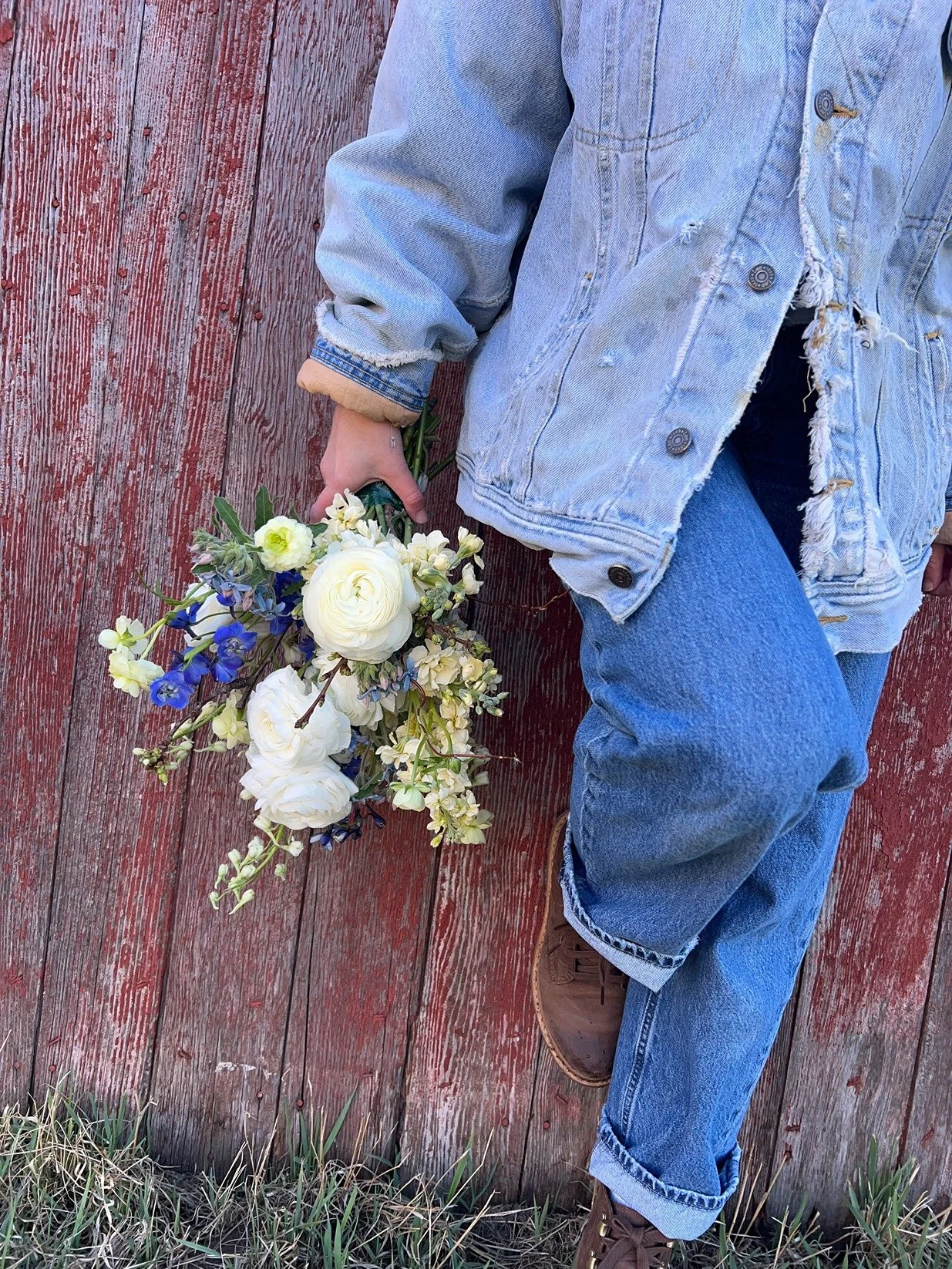 Person wearing a distressed denim jacket and blue jeans holding a bouquet of white, purple, and green flowers while standing on a red wooden surface outdoors.