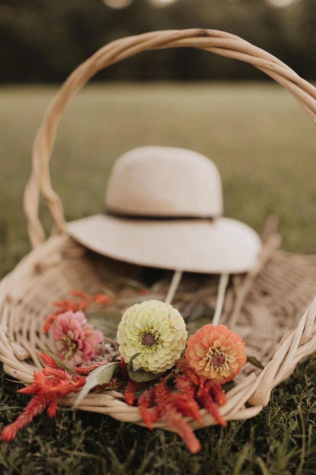 A wicker picnic basket on grass contains a wide-brimmed hat and a variety of colorful flowers, with a blurred background.