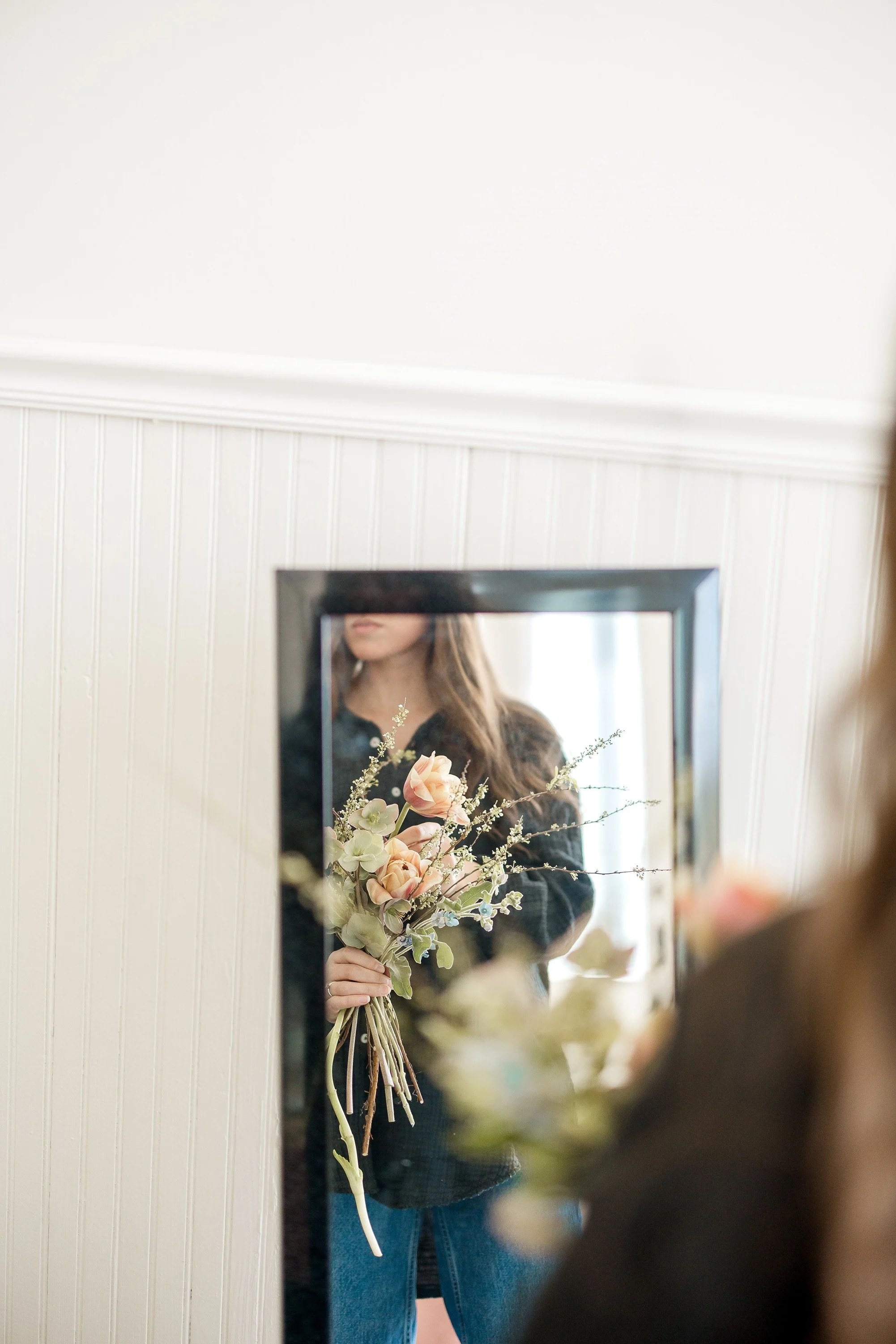 A person holding a bouquet of flowers is reflected in a mirror, with only partial visibility of their face and upper body.