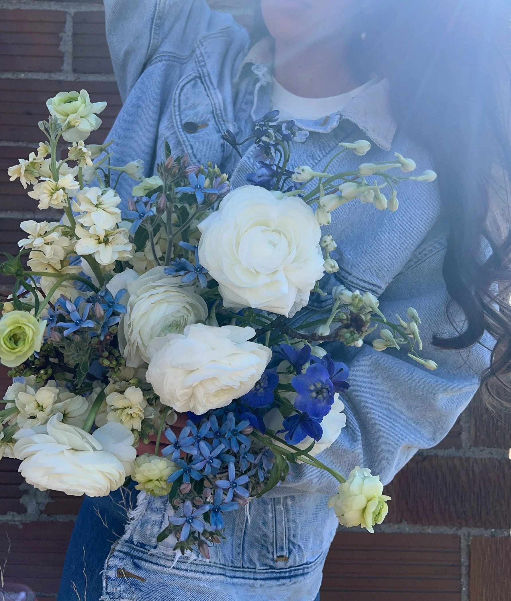 Person holding a bouquet of white and blue flowers, wearing a denim jacket and jeans, standing outdoors against a brick wall.