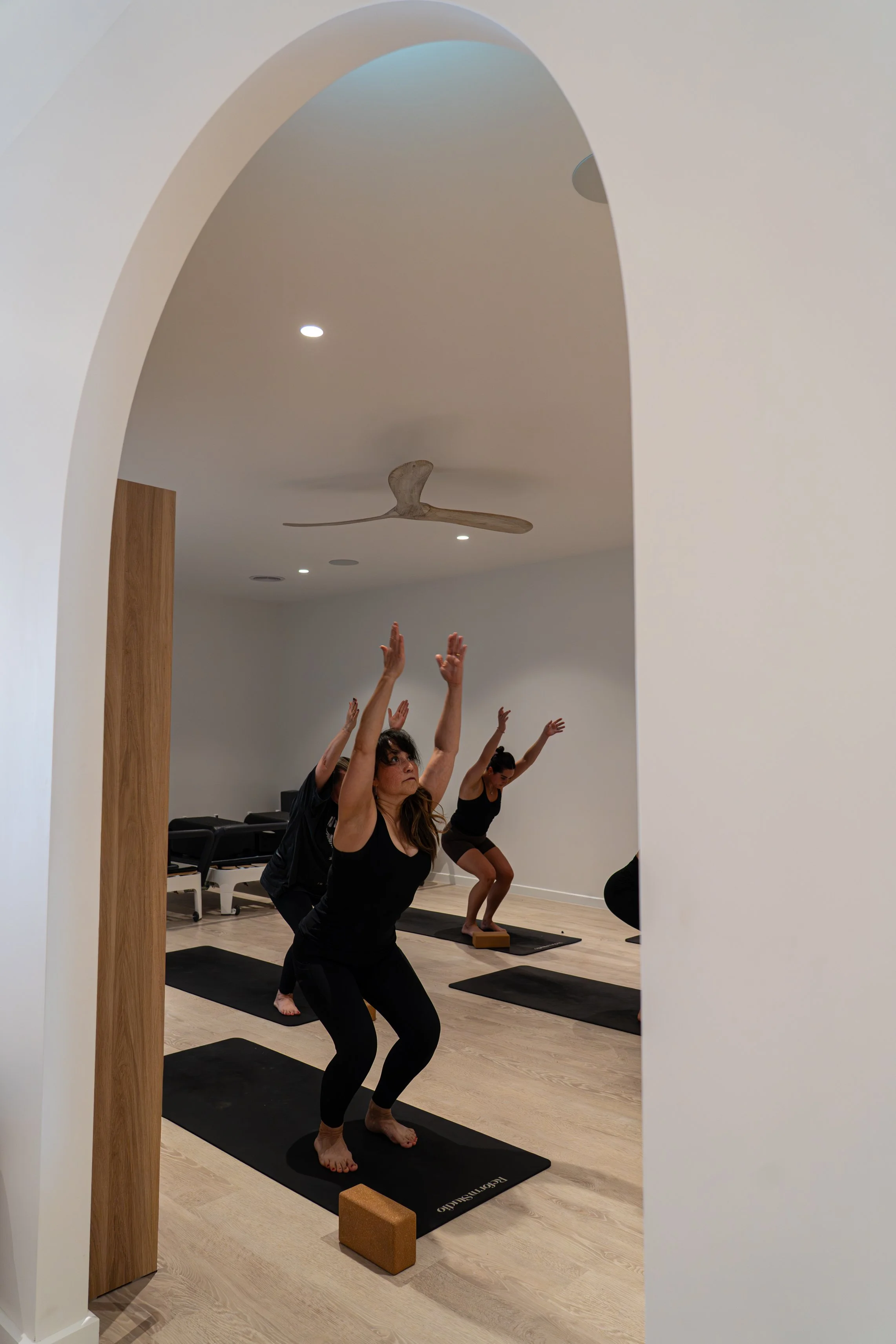 Women practicing yoga in a peaceful studio, raising arms in a pose, with yoga mats on the wooden floor and a ceiling fan overhead.