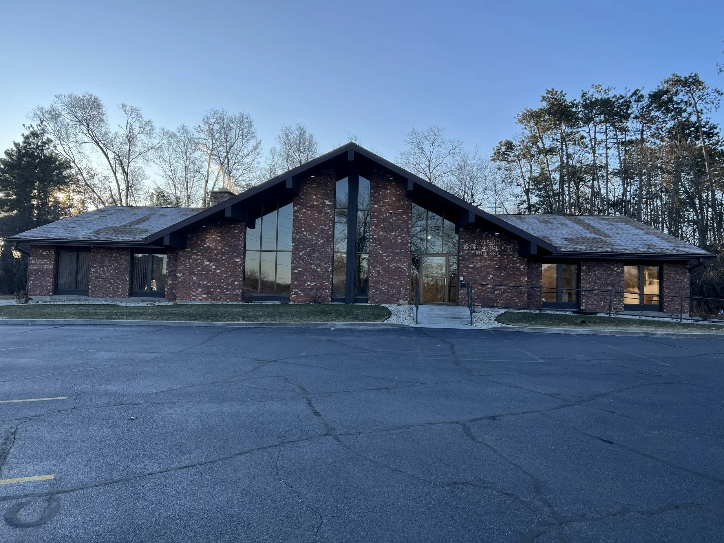Brick building with large glass windows and a sign reading 'Riverhill Professional Arts Building' on the front, surrounded by a parking lot and trees.