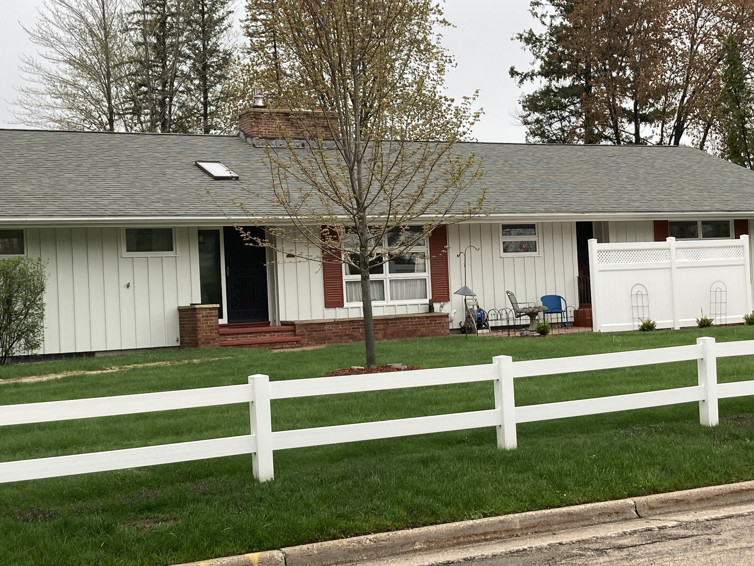 Single-story house with light siding, brick steps, and a front porch. There is a small tree in the yard, a white fence, and a seating area with chairs and a bird feeder.