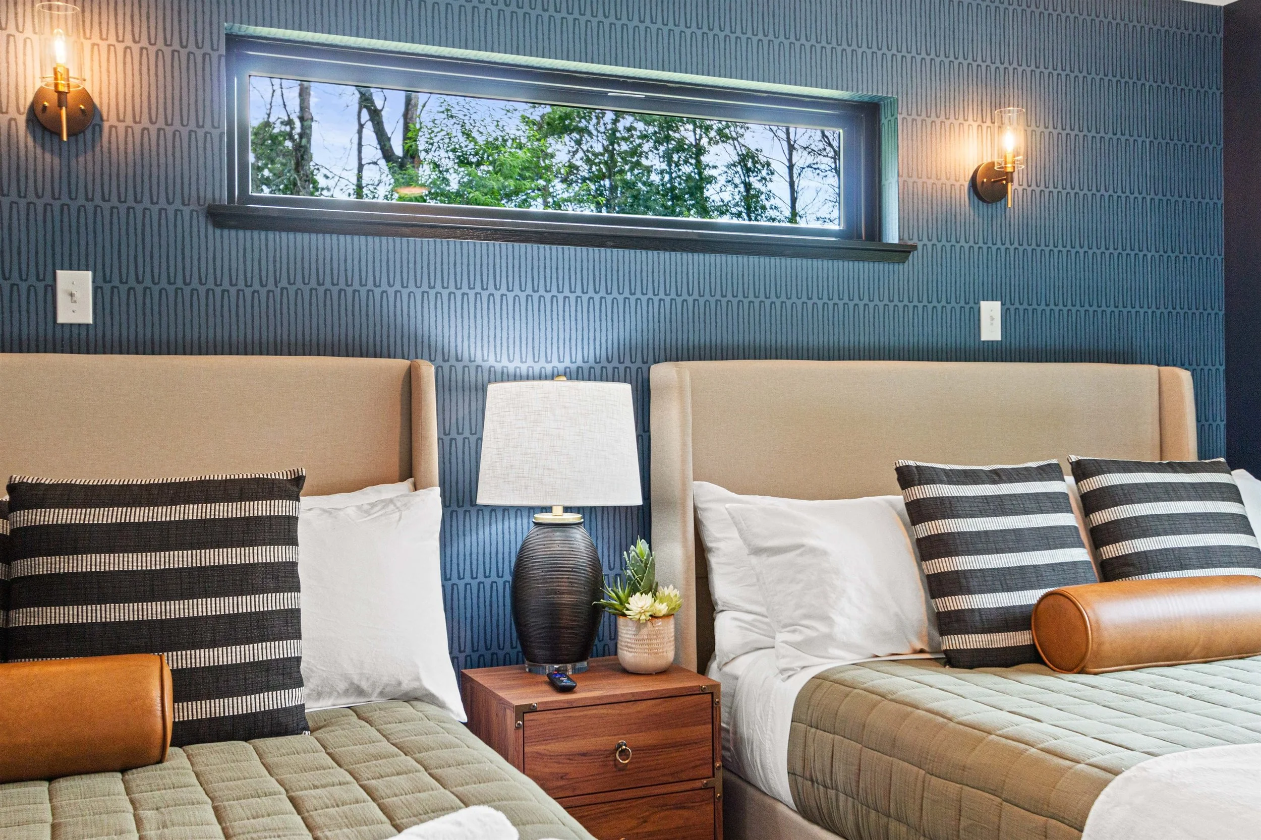 Two beds with beige headboards, striped black and white pillows, beige quilted blankets, and a bolstered pillow on each bed. A wooden nightstand between the beds holds a black table lamp with a white lampshade, a small potted plant, and a remote control. The wall behind is dark blue with textured patterns, and there are two wall sconces with visible filaments. Above is a narrow horizontal window showing trees and an overcast sky.