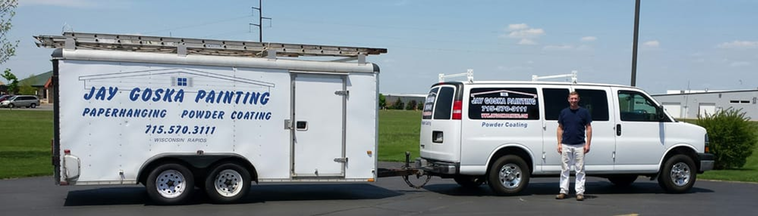A man stands next to a white van with a sign that reads 'Jay Goska Painting' parked on a paved area. The van is attached to a white trailer also with the same signage, and the trailer has a ladder on top. The background shows a grassy field and a few buildings under a blue sky with some clouds.