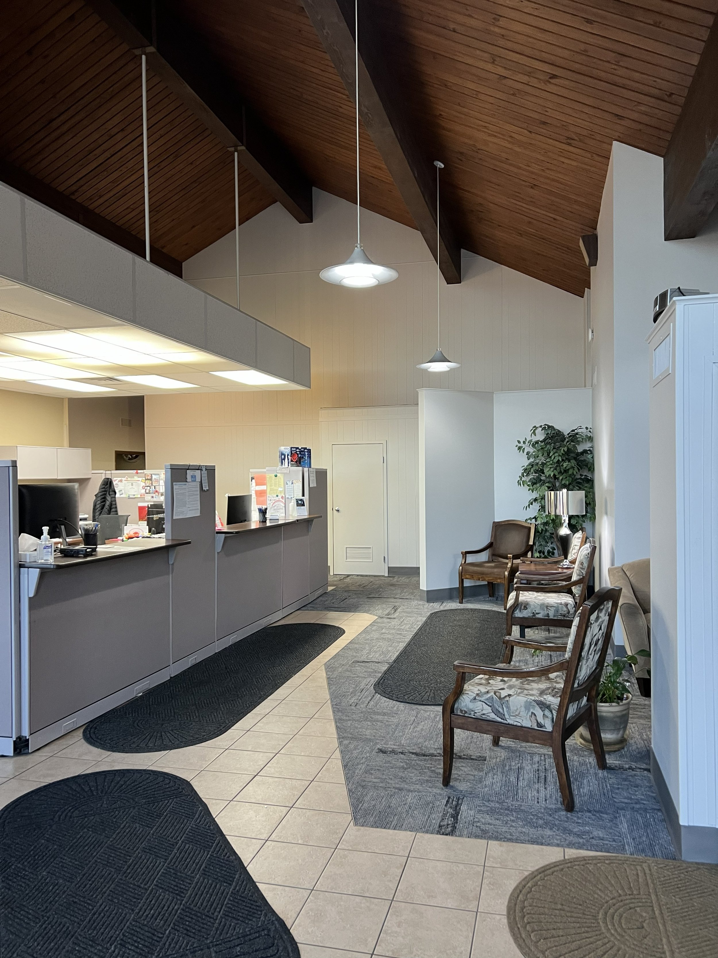 Interior of a reception area with a wooden vaulted ceiling, a reception desk with monitors and papers, and a seating area with several upholstered chairs, plants, and lamps.