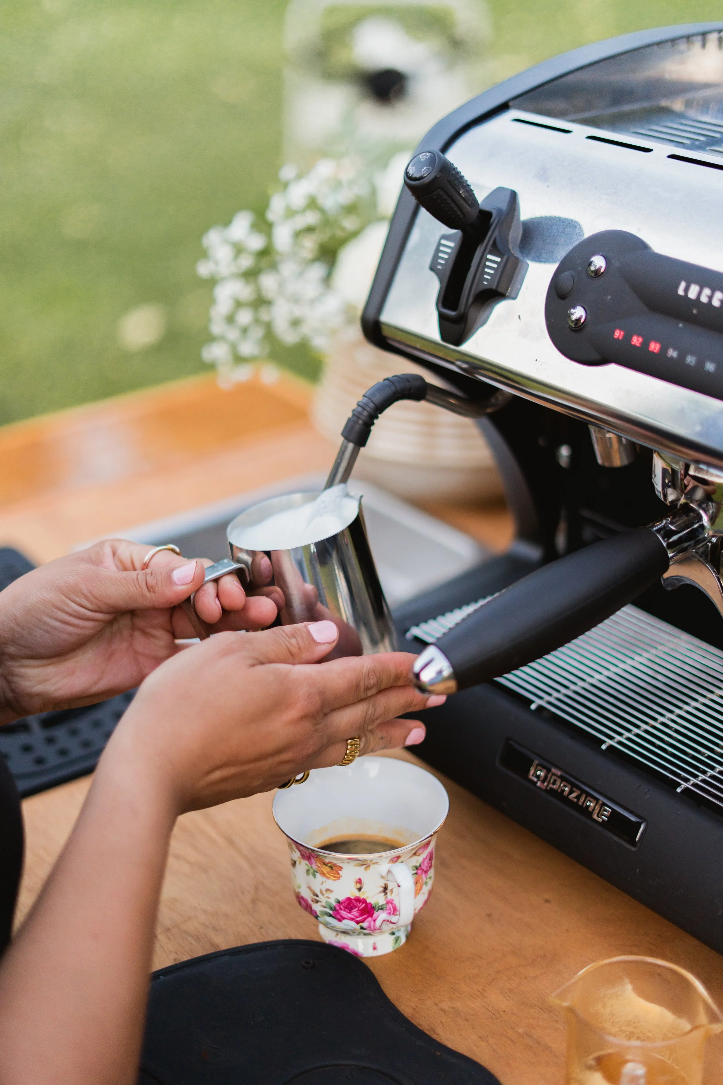 Person preparing a coffee drink using a coffee machine, pouring frothy milk into a floral mug.