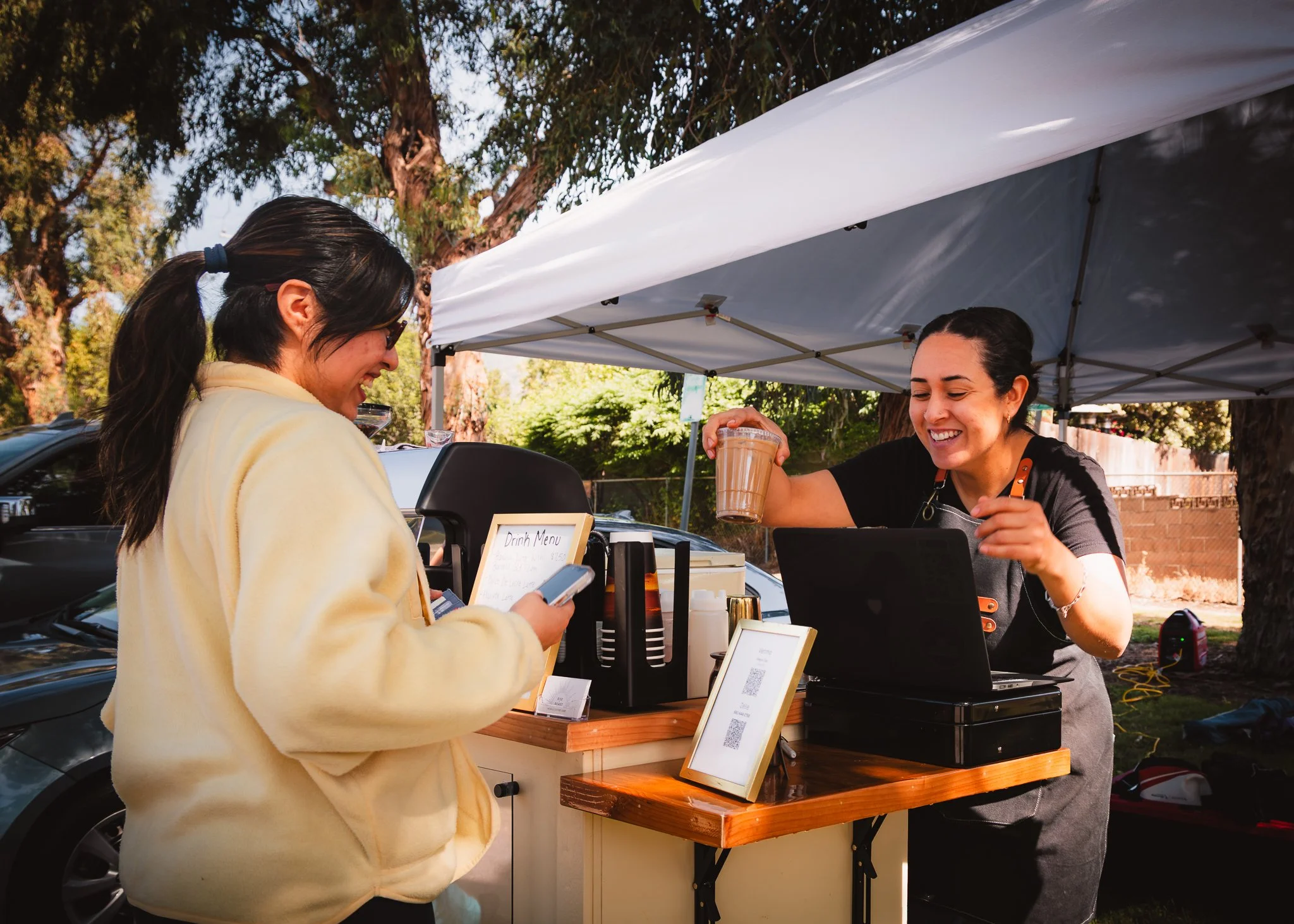 A woman at a food truck is handing a coffee drink to a customer, who is looking at her phone, under a blue canopy with trees in the background.
