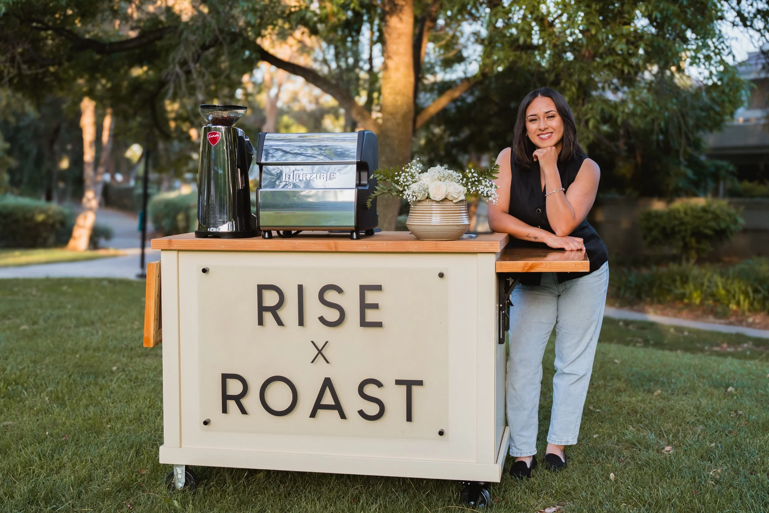 Smiling woman leaning on a food cart outdoors, with a sign that reads 'Rise x Roast,' a coffee grinder, and a flower vase, surrounded by trees.