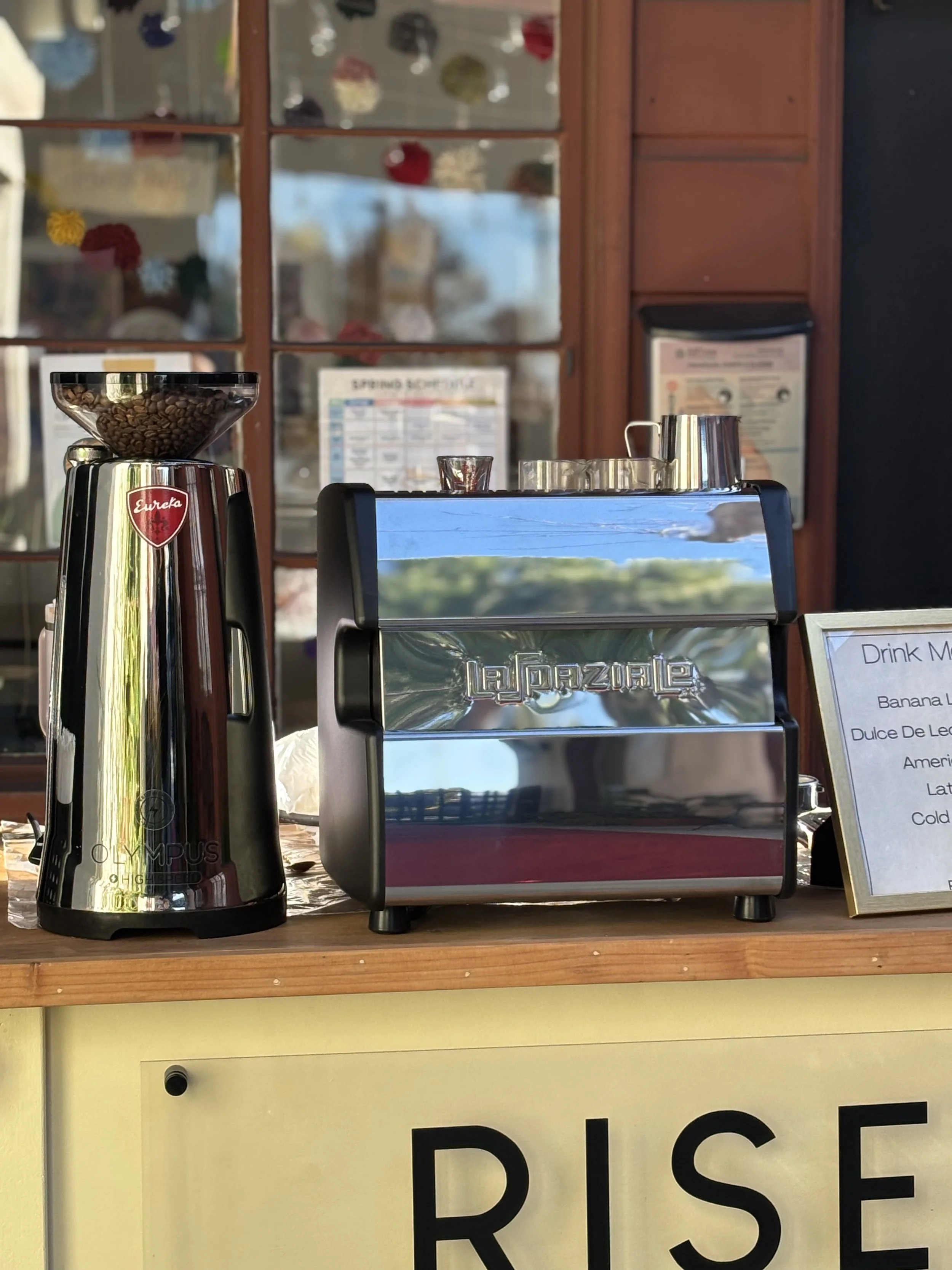 Coffee grinder and  espresso machine on a wooden counter at a café, with a menu sign and a window in the background.