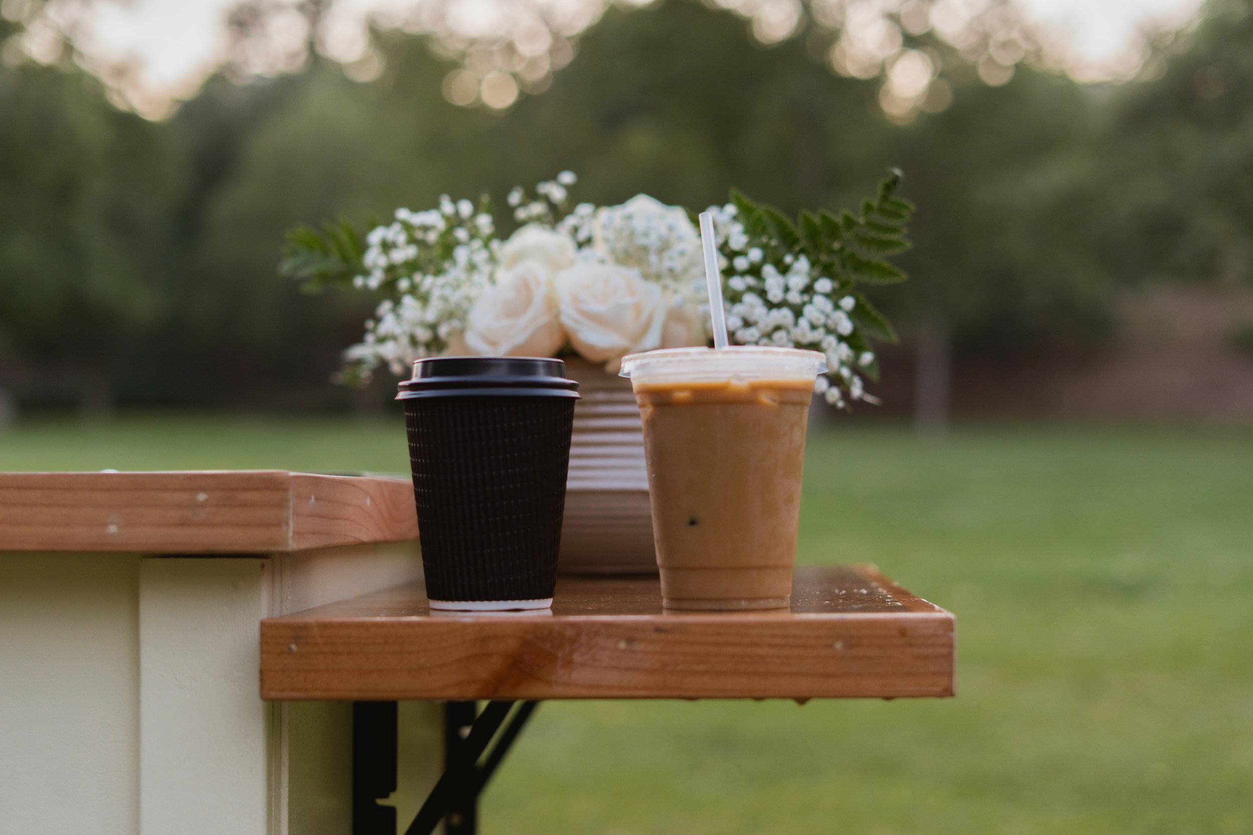 Two takeaway coffee cups and a plastic cup with iced coffee or tea on a wooden table outdoors, with a floral arrangement of white roses and baby's breath in the background, blurred greenery and trees.