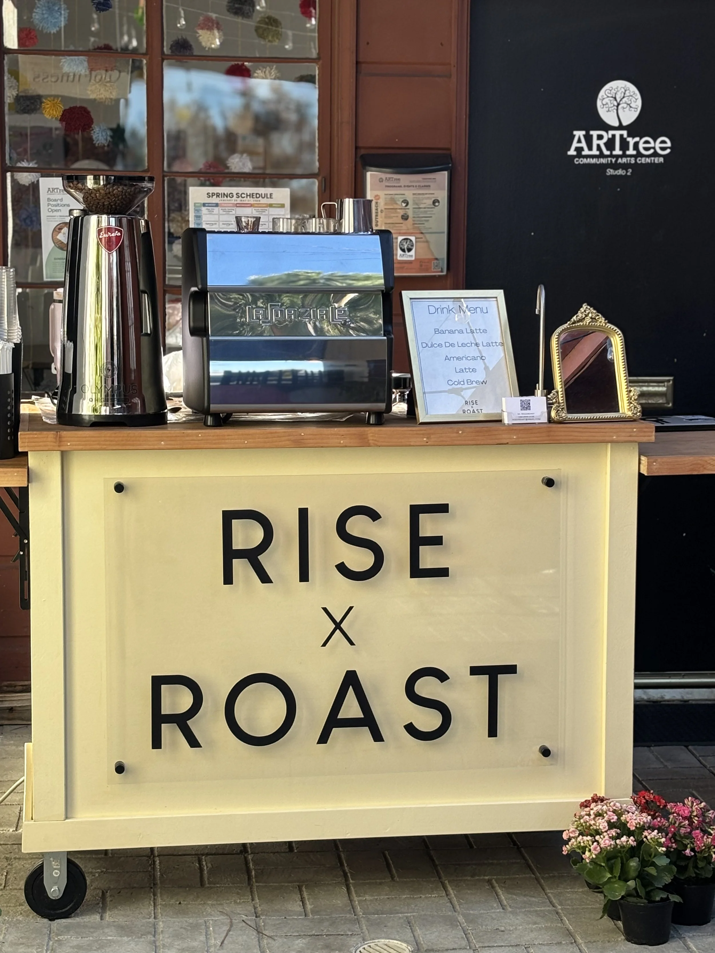 A coffee stand with a sign that says 'RISE x ROAST', a coffee grinder, an espresso machine, a drink menu, and a small decorative mirror. There are flowers in a pot at the bottom right corner.