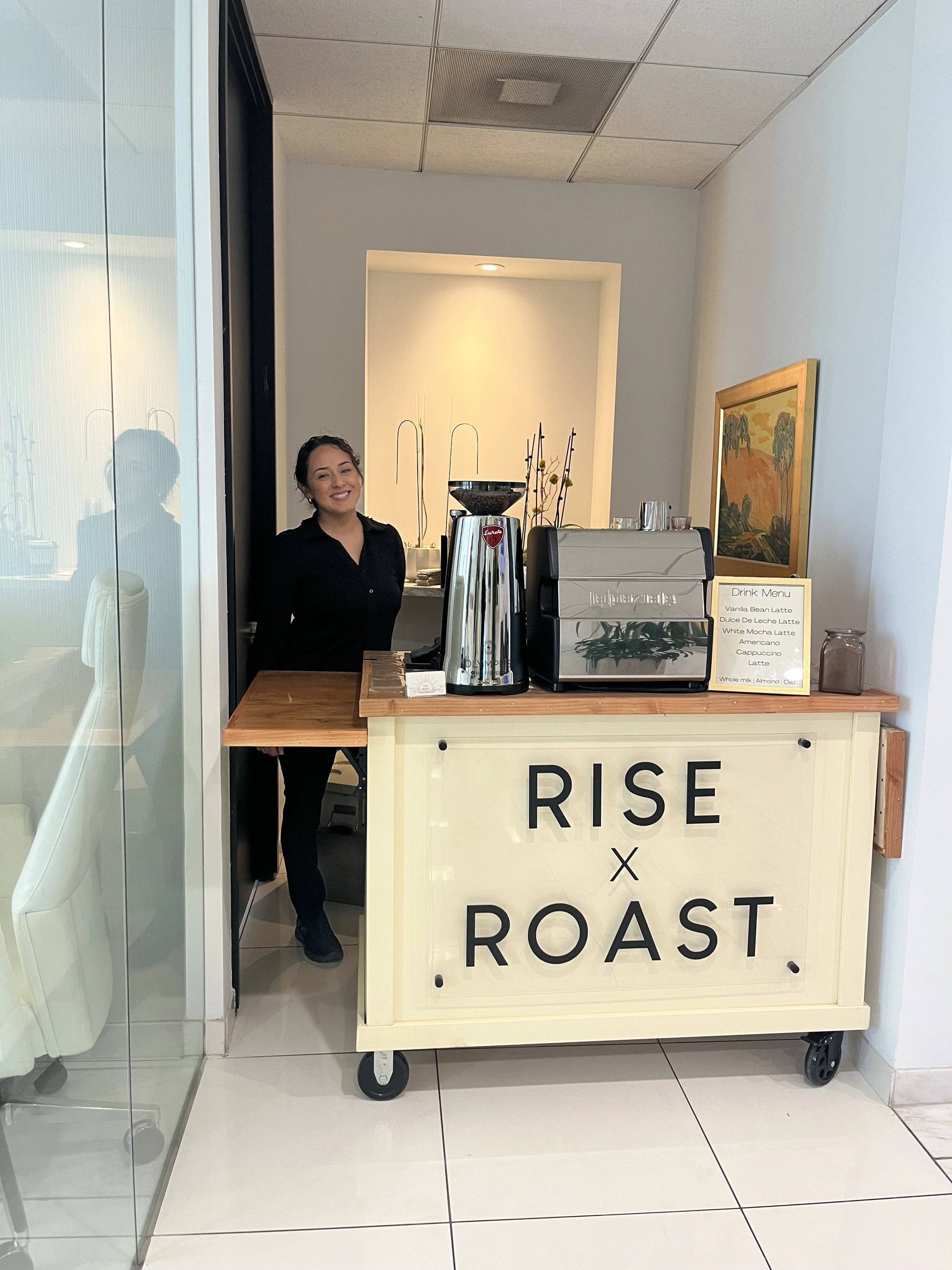 A woman standing behind a coffee cart with a sign that reads 'Rise x Roast.' The cart has a coffee machine, a large coffee grinder, and a drink menu. The woman is smiling and wearing a black shirt, inside a modern building.