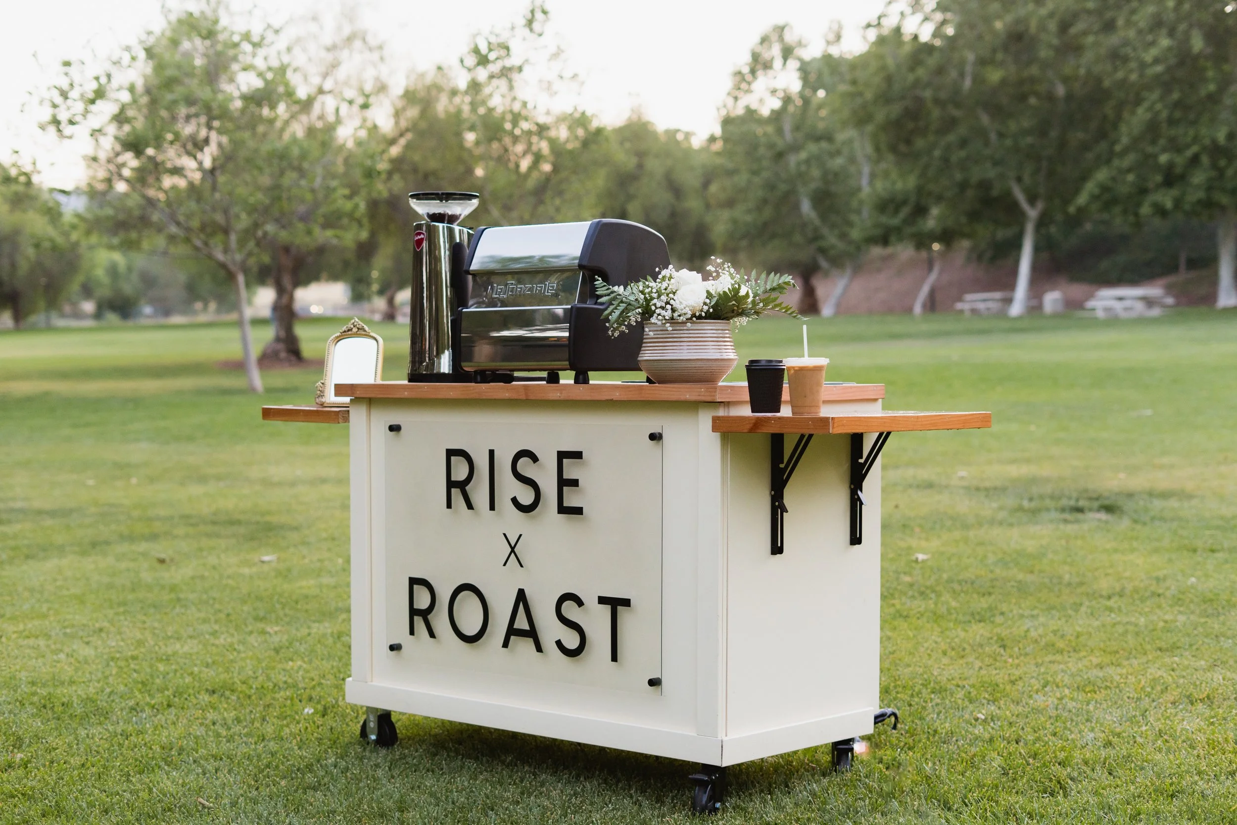 A mobile coffee stand with a sign that reads 'Rise x Roast' set up in a park with trees in the background. The stand has a coffee machine, flowers in a vase, and two cups.