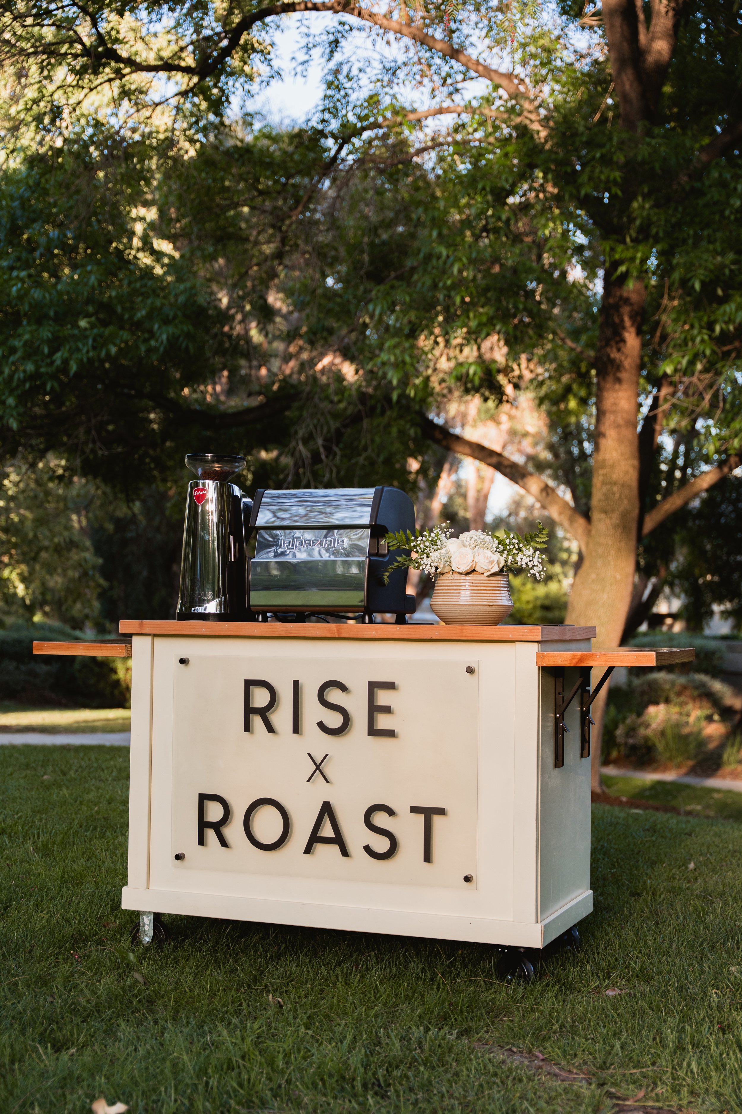 A table displaying coffee equipment and flowers in a park setting with trees and sunlight in the background. The table has a sign that says "Rise x Roast."