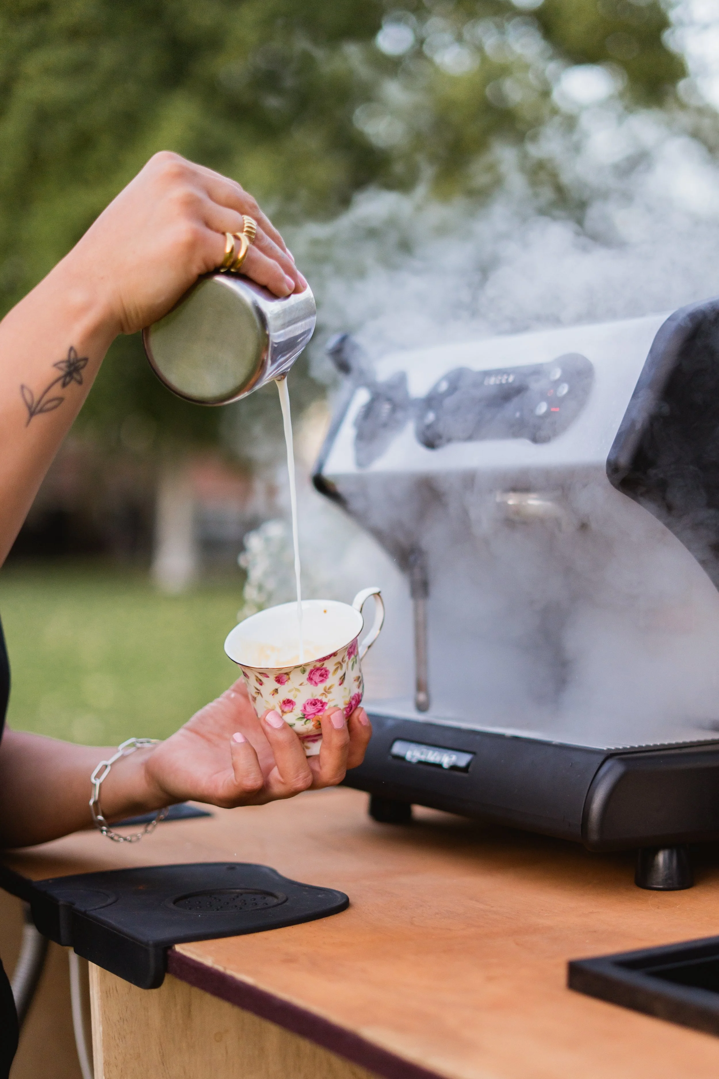 Person pouring coffee into a floral teacup held by another person in front of a coffee machine outdoors.