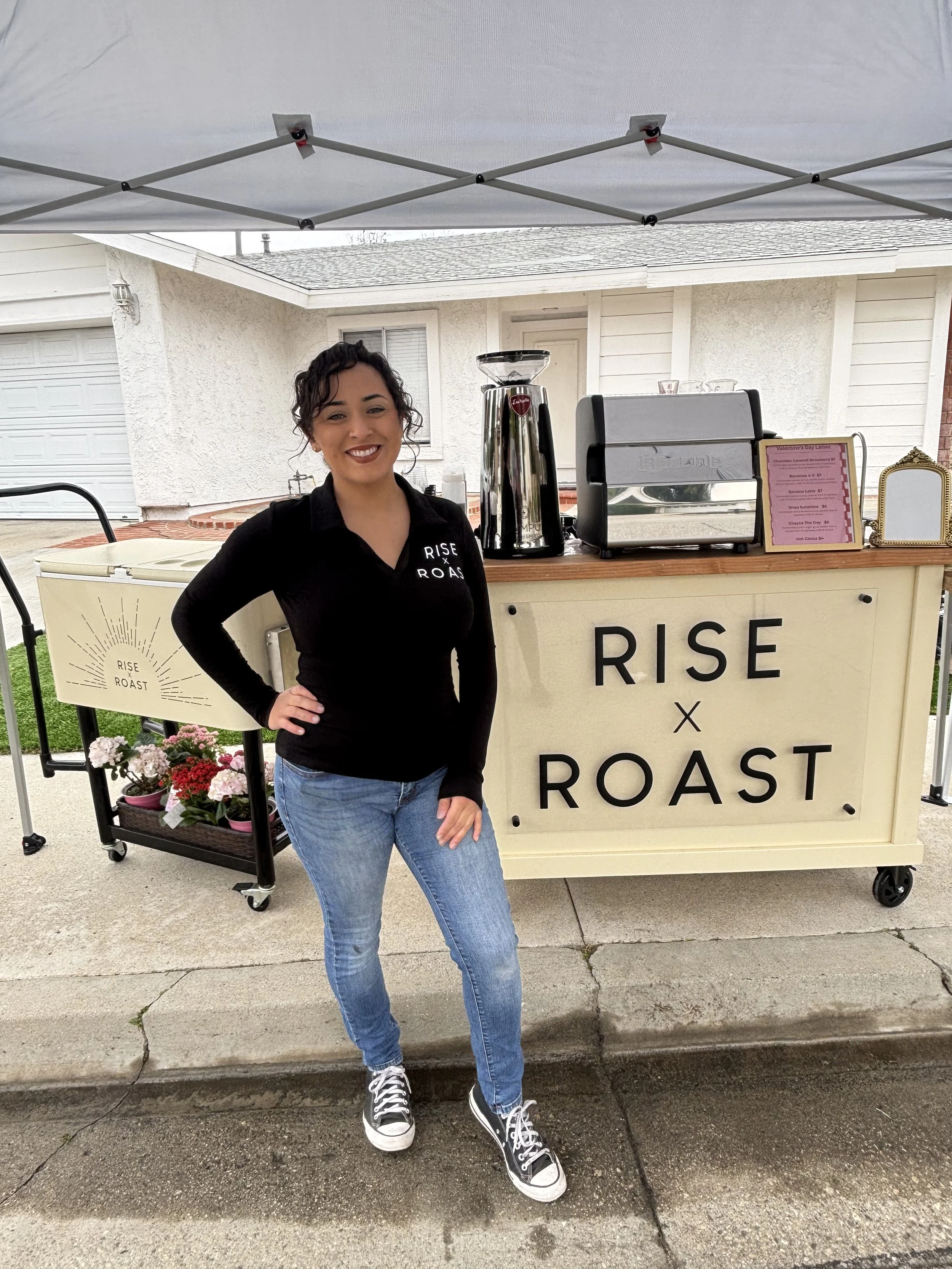 A woman standing in front of a coffee stand that has the words 'Rise x Roast' on it. She is smiling, dressed in a black sweater and blue jeans, with a hand on her hip.