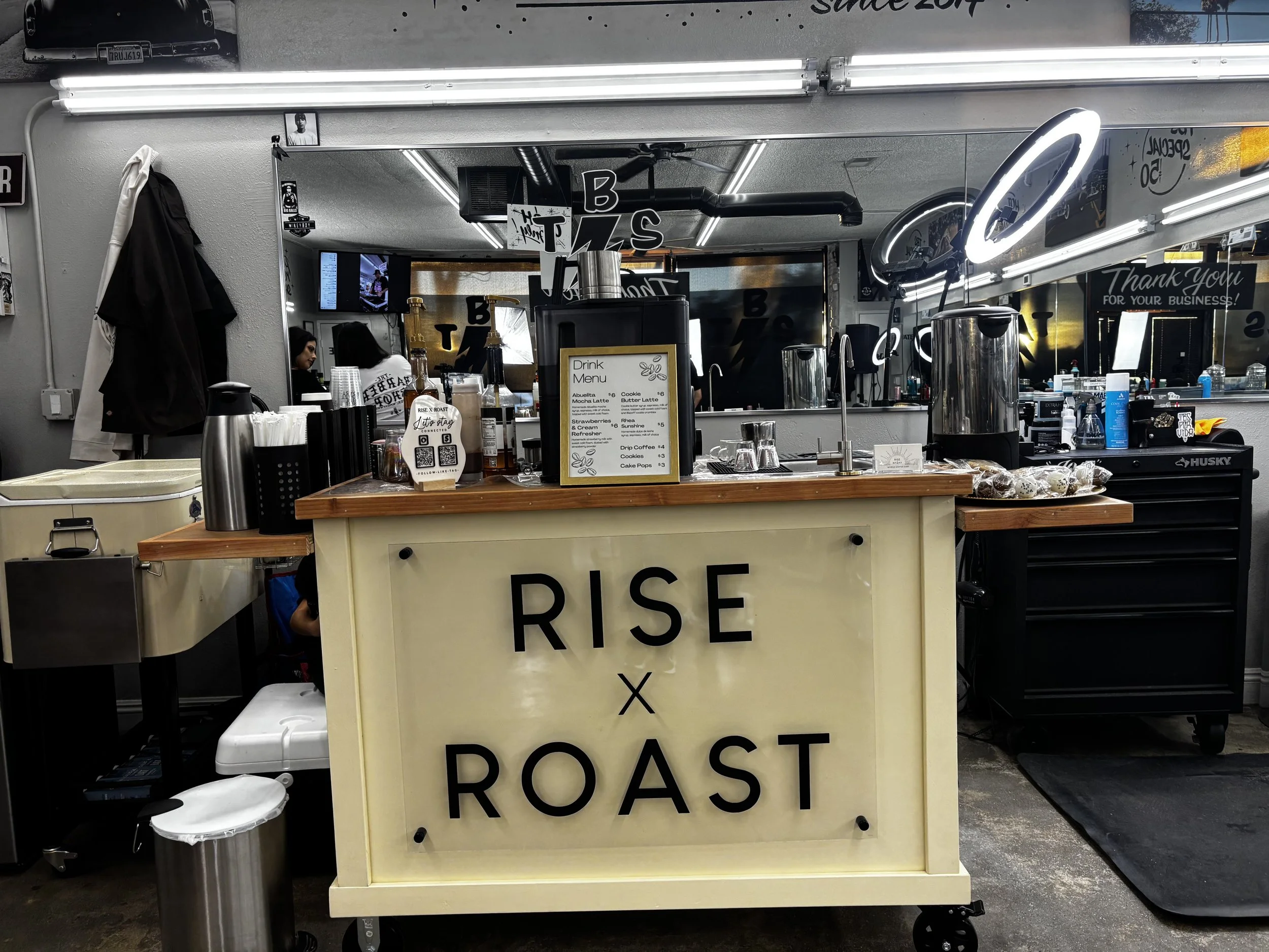 A coffee stand with a sign that says "RISE x ROAST" in black letters on a yellow background. The stand has coffee cups, bottles, a drink menu, and equipment on top. There is a mirror behind the stand with partial reflections of the interior of a coff