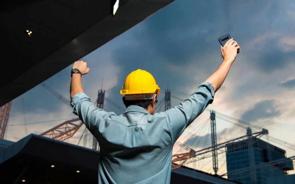 A construction worker wearing a yellow hard hat and light blue shirt taking a selfie with a smartphone against a cityscape with cranes and buildings during sunset.