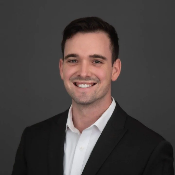 A young man in a white shirt and black blazer smiling against a dark gray background.