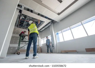 Construction workers installing or inspecting ceiling panels in a large, well-lit room with windows.