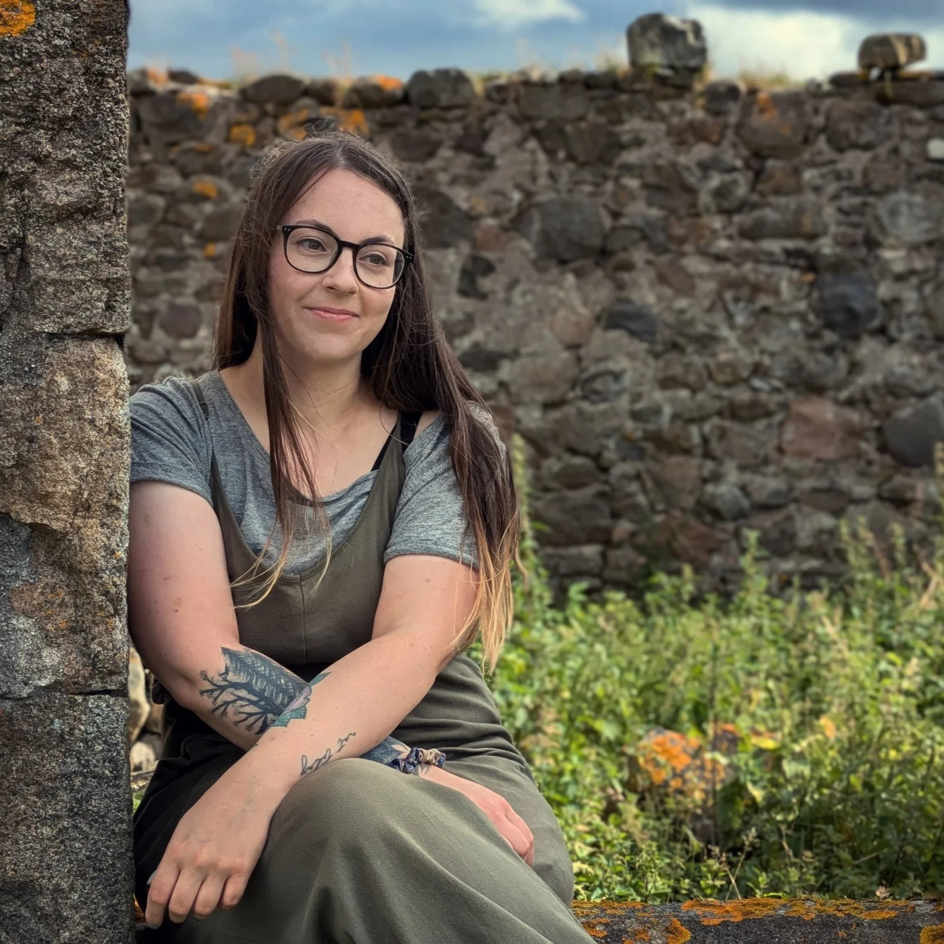 A young woman with long dark hair, glasses, and tattoos on her arm, sitting outdoors against a stone wall with green foliage in the background.