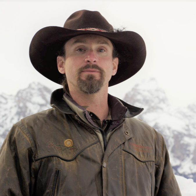 A man wearing a cowboy hat and brown leather jacket standing outdoors with snow-covered mountains in the background.
