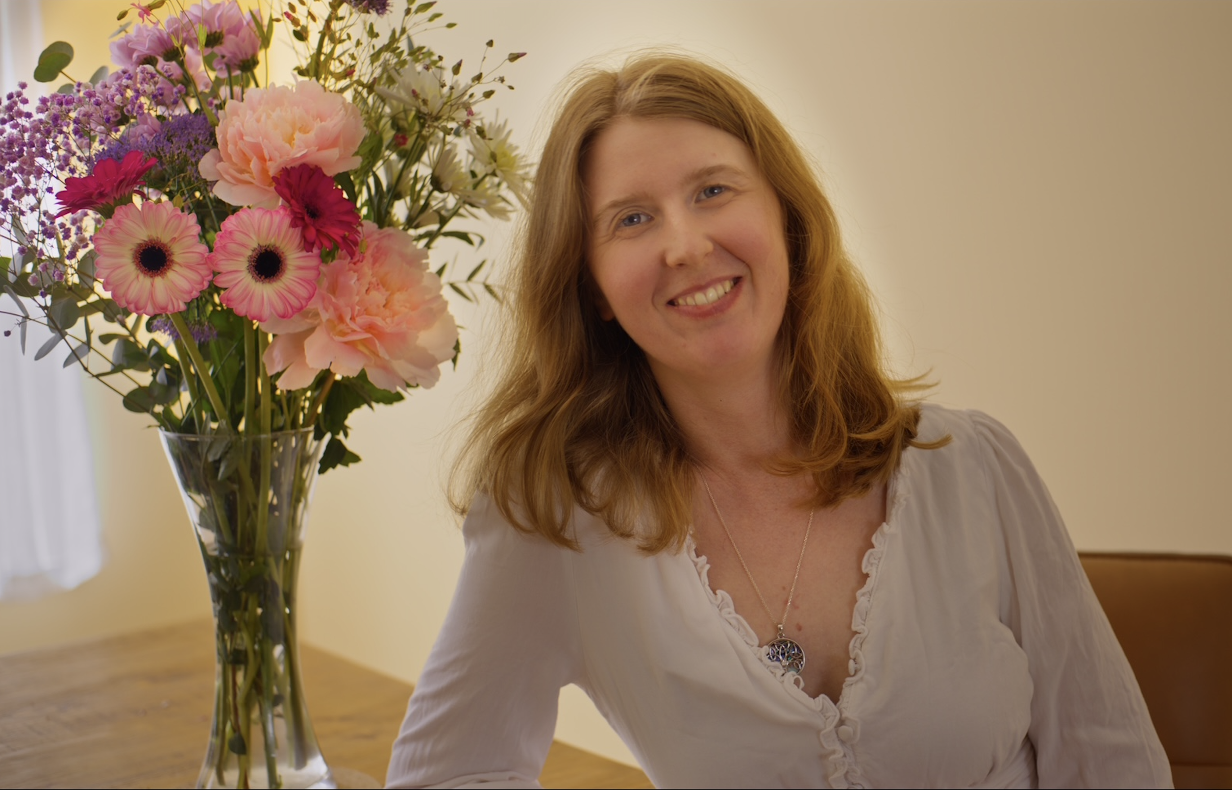 A smiling woman with red hair sitting at a wooden table, next to a large bouquet of pink, purple, and white flowers in a glass vase, in a room with beige walls.