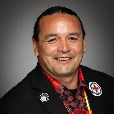 A smiling man with dark hair, wearing a black blazer with buttons and a patterned red and black shirt, against a gray background.