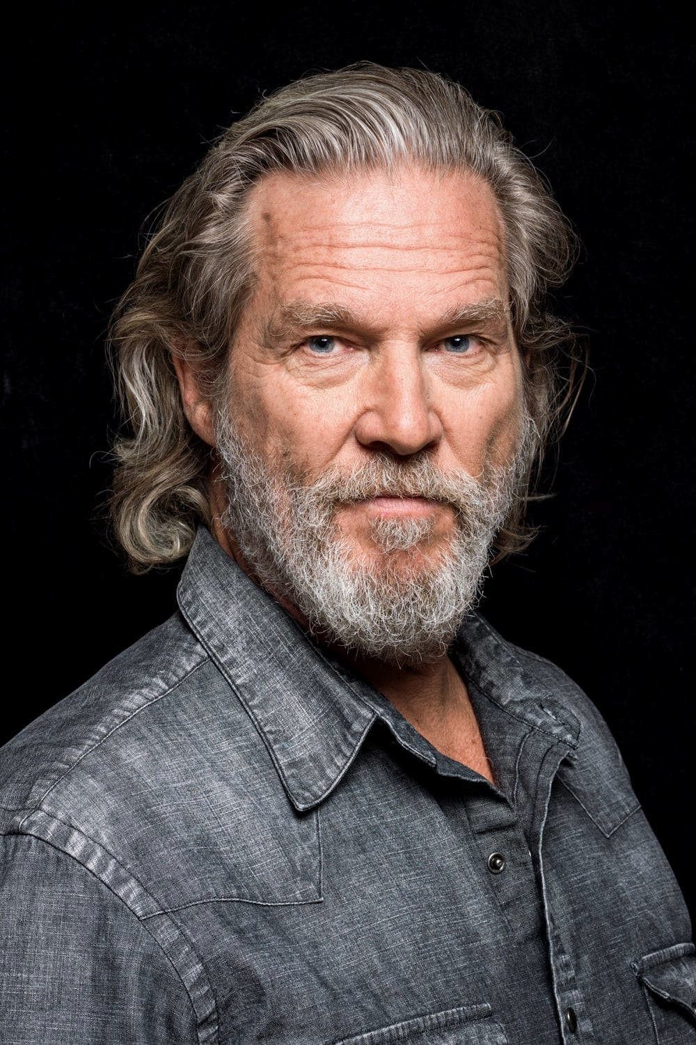 Close-up portrait of an older man with gray hair, blue eyes, and a beard, wearing a gray button-up shirt against a black background.