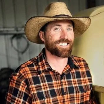 A man with a beard wearing a cowboy hat and a plaid shirt, smiling in a rustic setting.