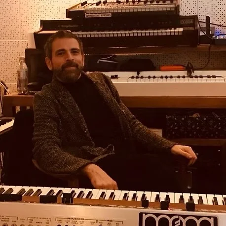 Man sitting in a recording studio with electronic music equipment including keyboards and synthesizers.