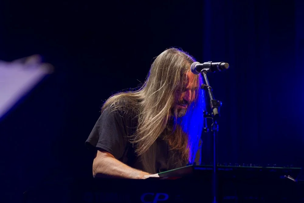 A long-haired man performing on stage with a keyboard and microphone, under purple lighting.