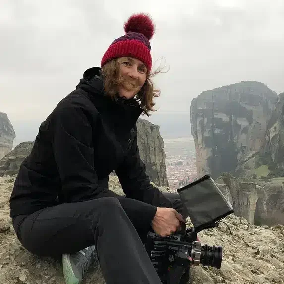Woman sitting on rocky terrain with a camera, wearing a black jacket and a red knit hat with a pom-pom, mountains and a cloudy sky in the background.