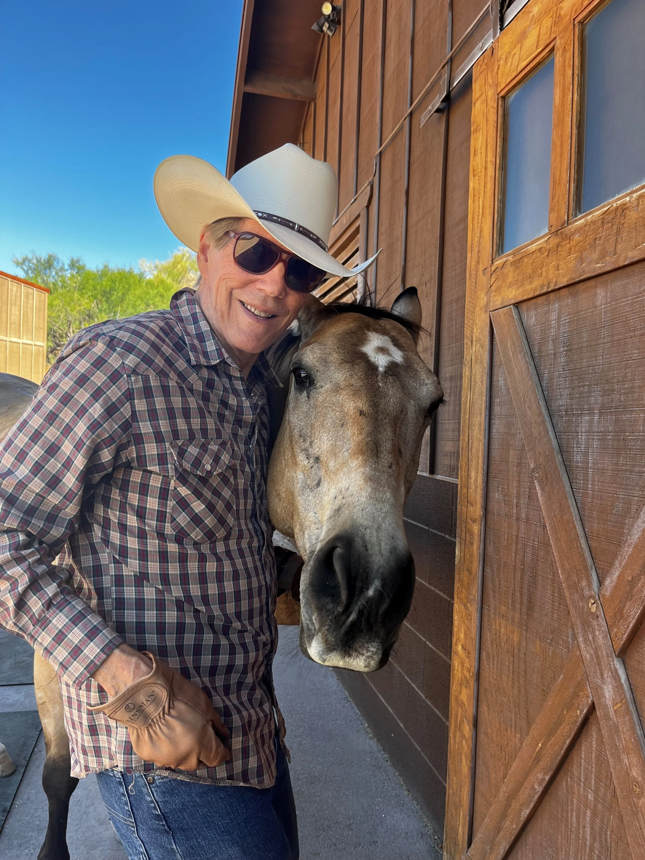 A man wearing a cowboy hat, sunglasses, and a plaid shirt is smiling and hugging a horse's face, which is next to a wooden barn door.
