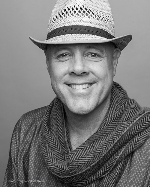 A smiling man wearing a light-colored fedora hat with a dark band, a patterned shirt, and a scarf. The image is in black and white.