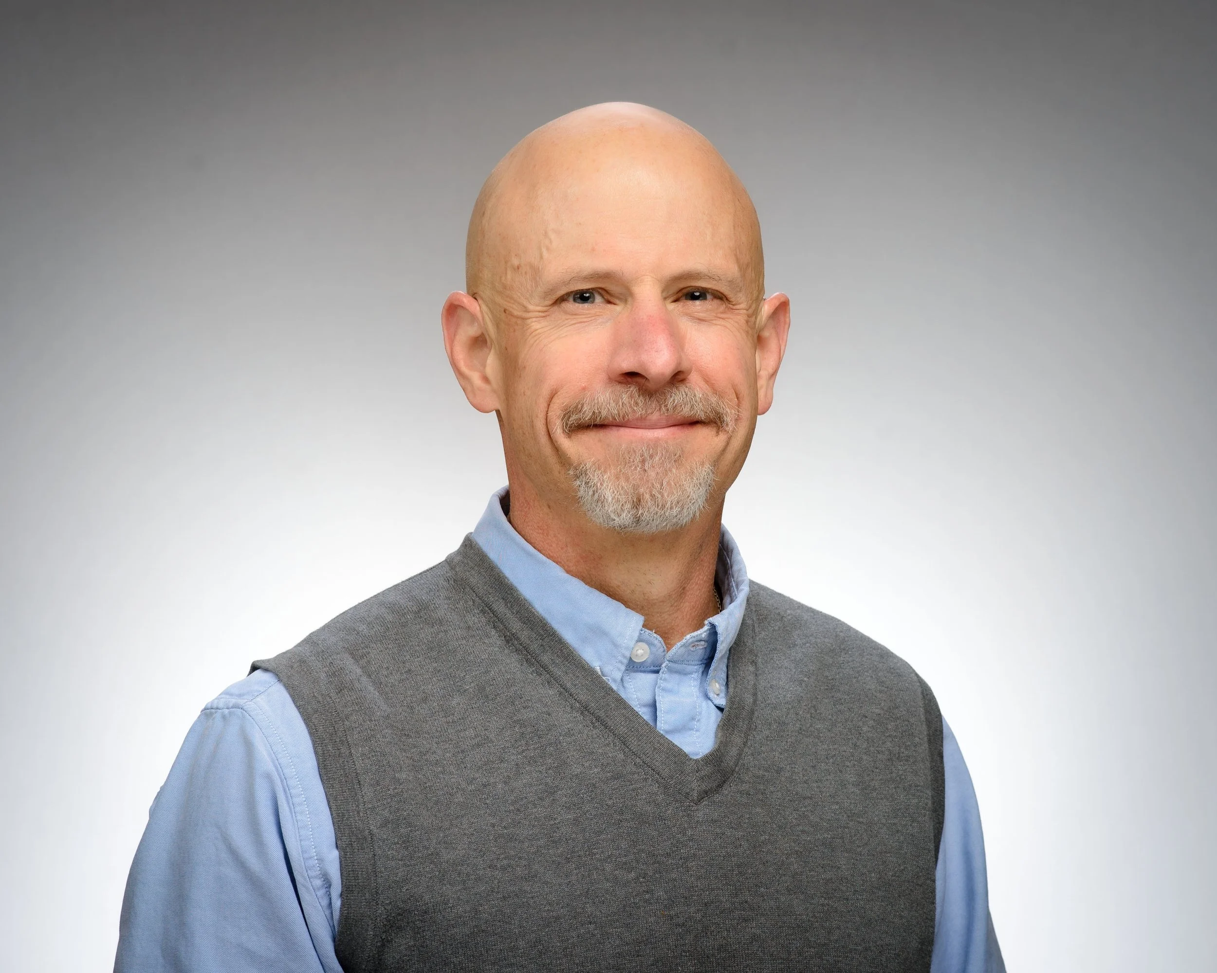 A smiling middle-aged man with a bald head and gray goatee, wearing a blue shirt and gray sweater vest, against a plain gray background.