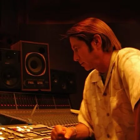 A man with brown hair and a yellow shirt sitting in a recording studio, working at a mixing console with speakers behind him.