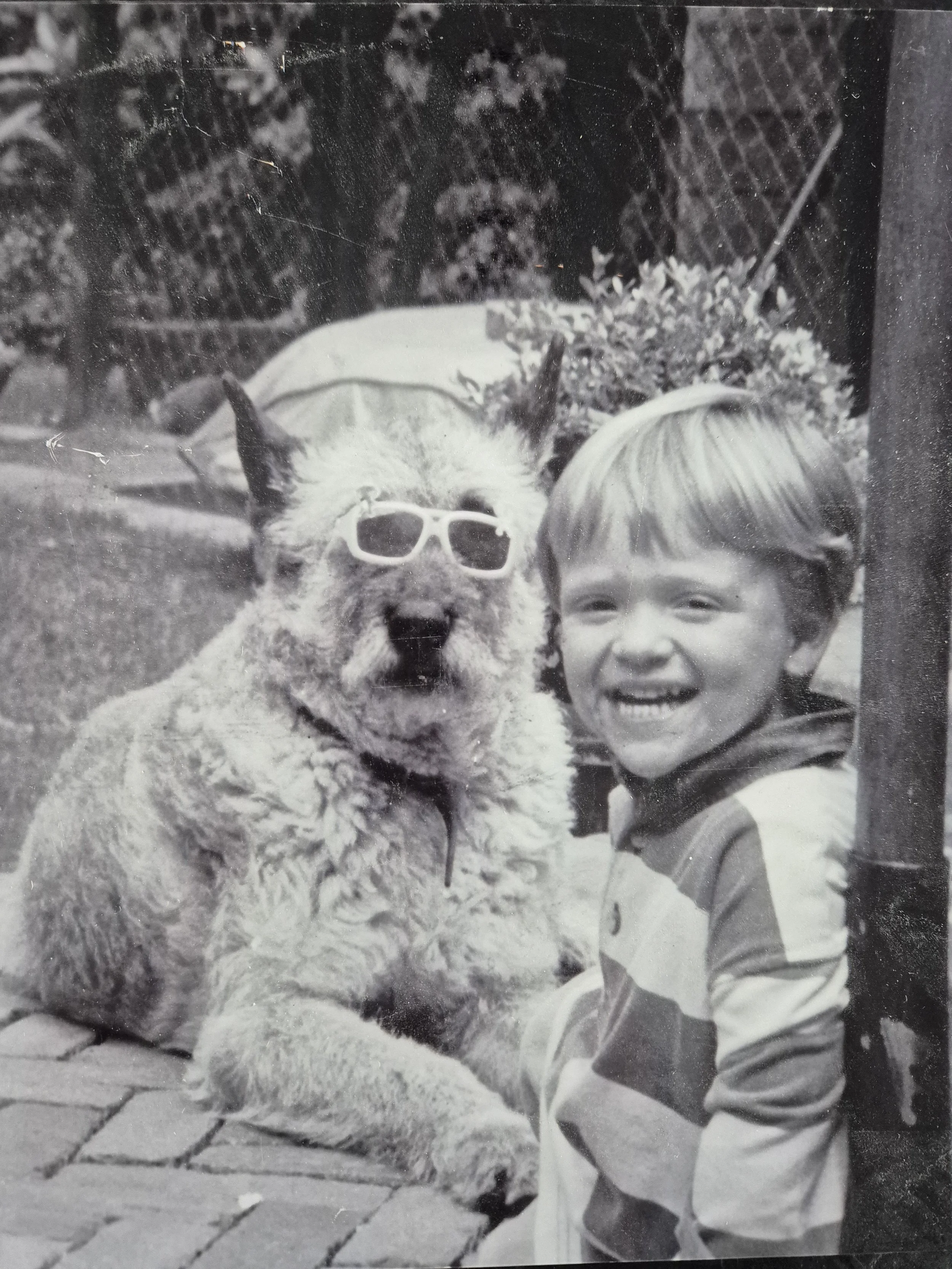 Een zwart-wit foto van een jongen met blond haar die lacht, naast een hond met zonnebril, in een tuin met een metalen hek op de achtergrond.