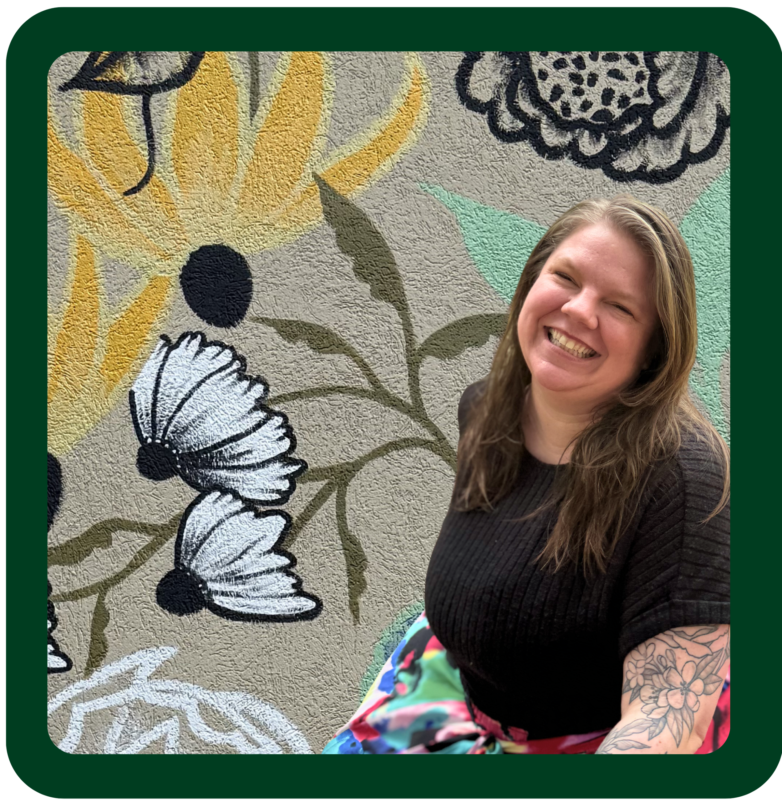 Photo of Miranda Hosbein LMFT in a black top and rainbow skirt  smiling warmly. Miranda is standing in front of a local Kalamazoo mural that depicts different flowers behind her. Mural was painted by artist Paige Rochefort