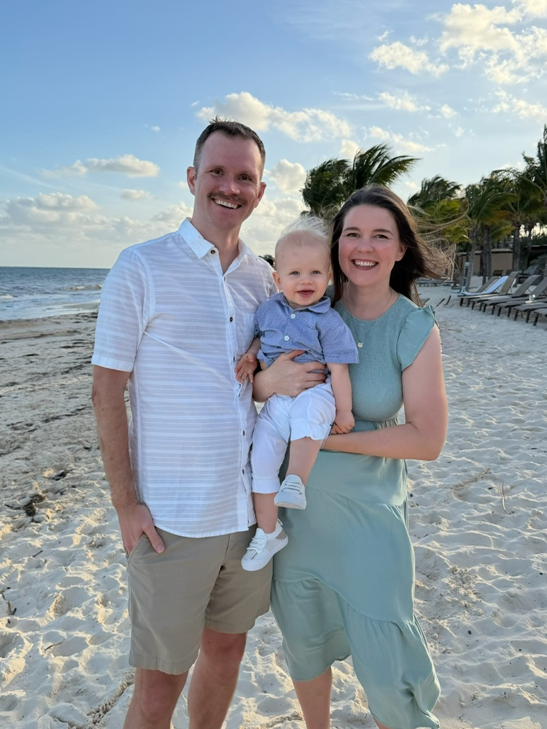 Jay with his wife and young son on vacation on a beach, with palm trees, lounge chairs, and the ocean in the background, during sunset.