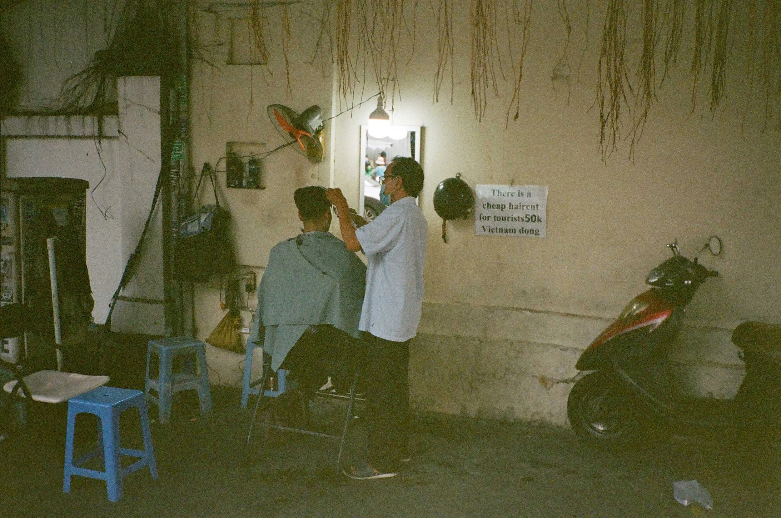 A man giving a haircut on the sidewalk under a single light and small mirror.