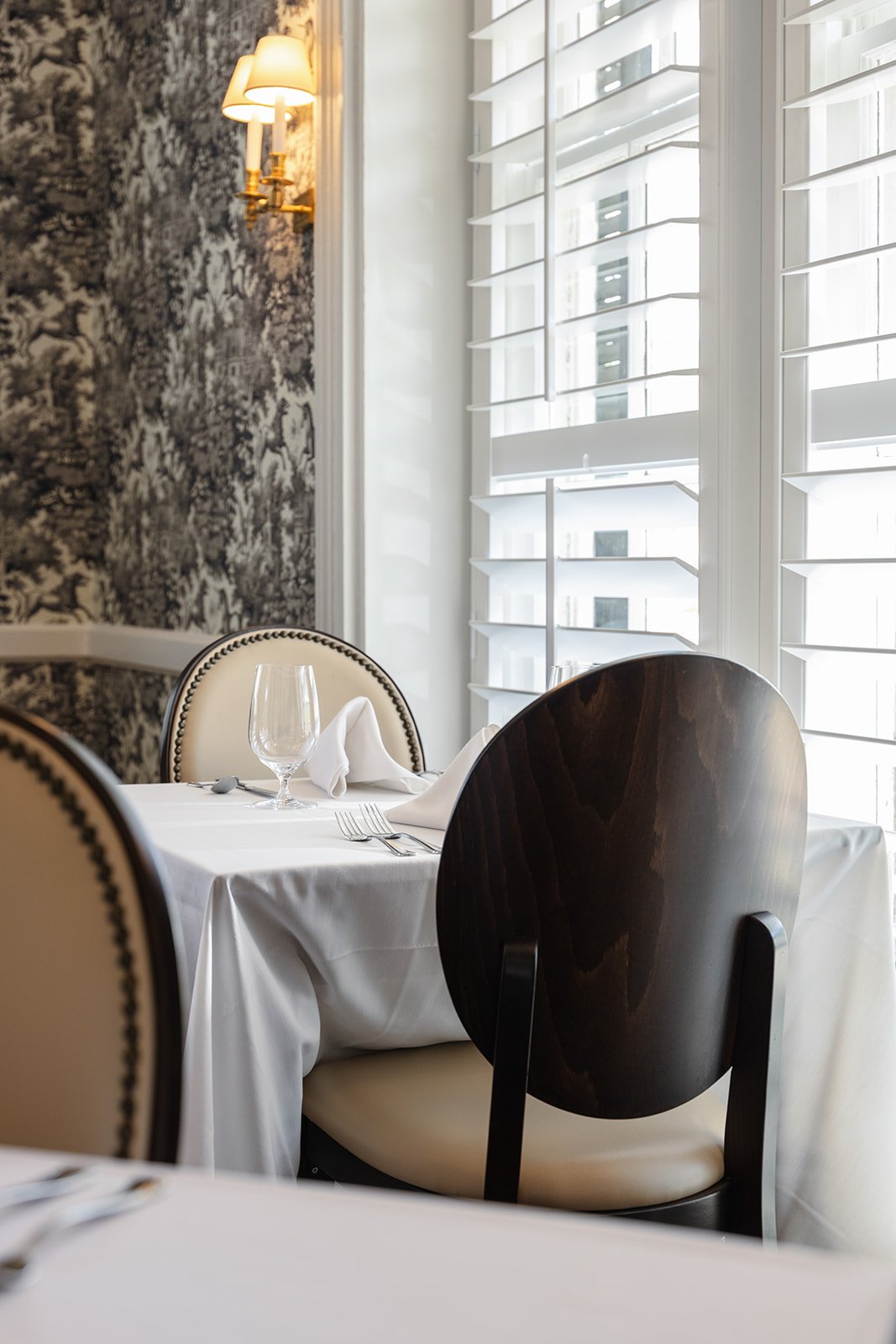 Carlisle's Restaurant - Table for 2 - Elegant restaurant table set with white tablecloth, a wine glass, a folded napkin, and cutlery, near a window with white shutters, and a wall with a black and white textured mural.